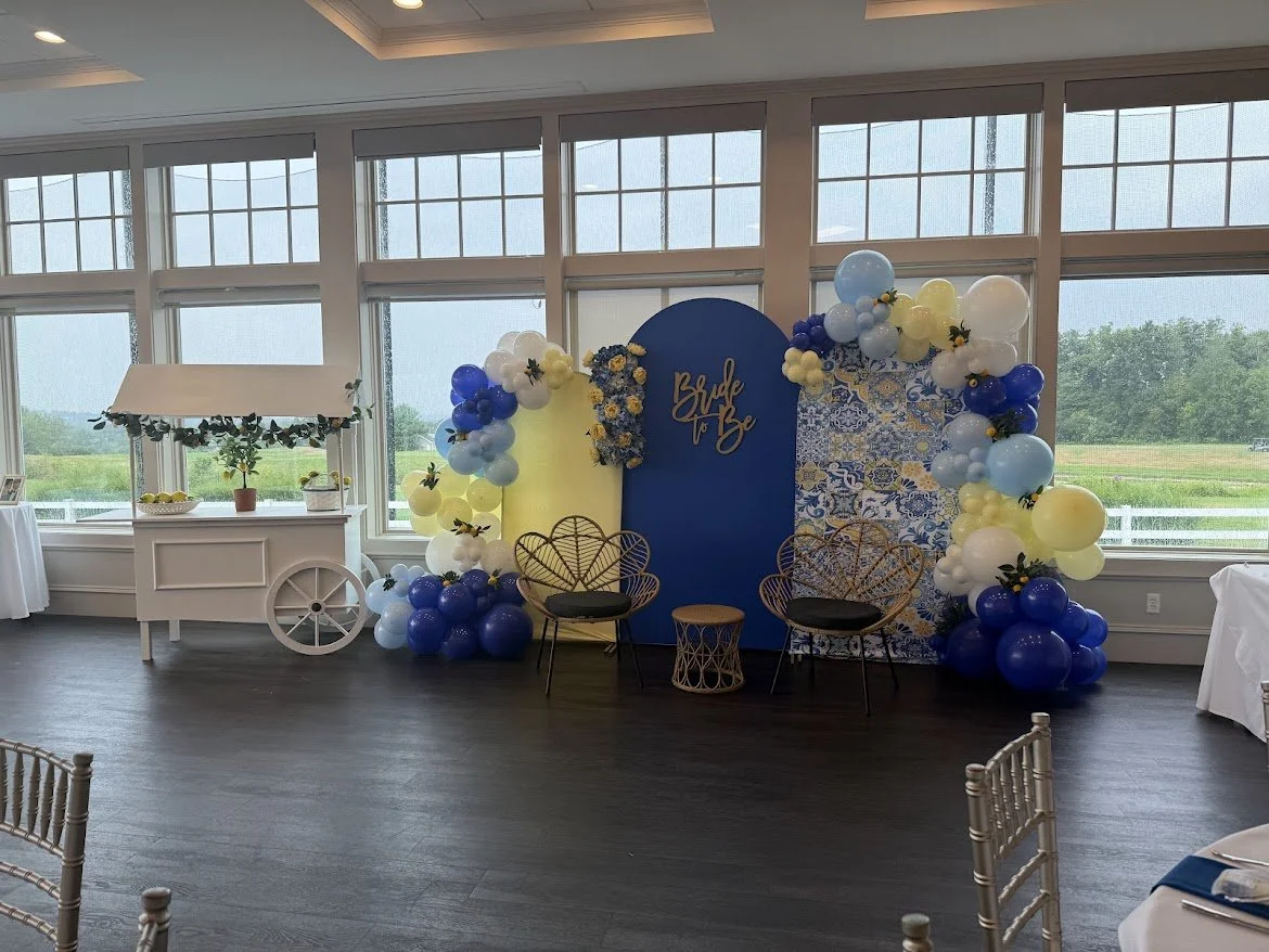 Decorative backdrop with balloons and a sign that reads 'Bride to Be' at a bridal shower event in a bright room with large windows and views of the outdoors.