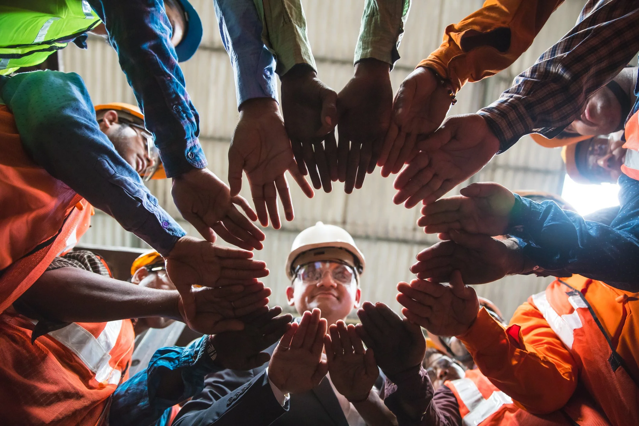 A group of diverse workers wearing safety helmets and vests forming a circle with their hands together, looking down at the center.