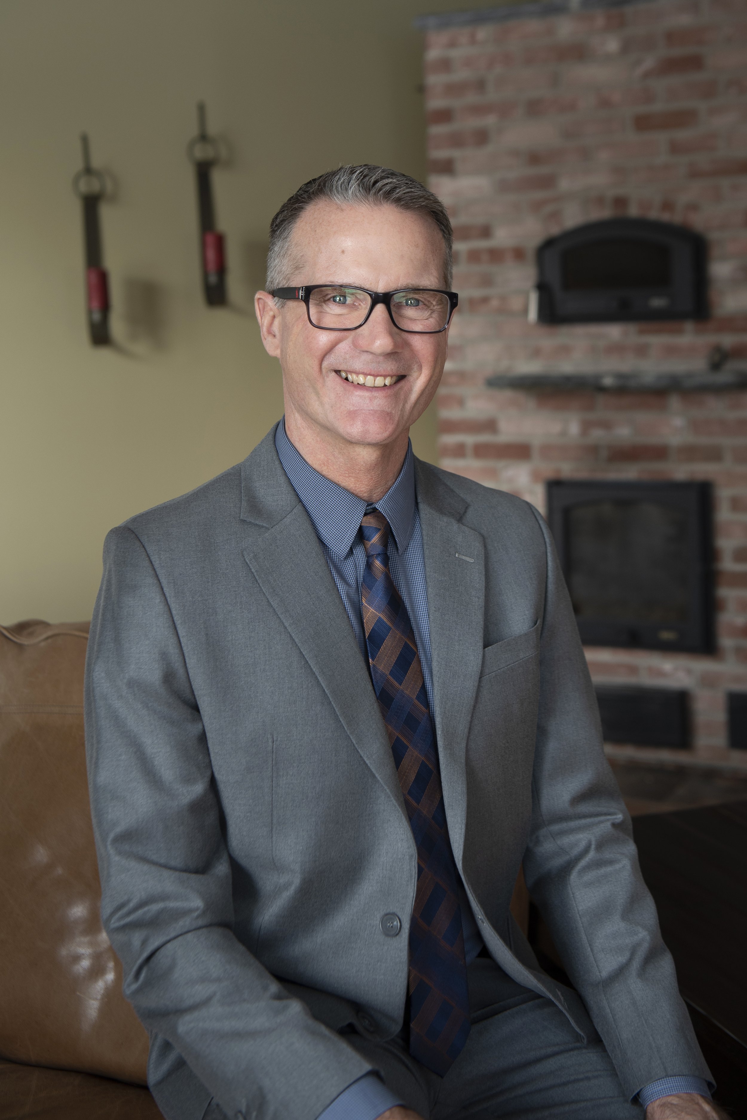 A man in a gray suit, blue shirt, and patterned tie, wearing glasses, sitting in a living room with a brick fireplace in the background.