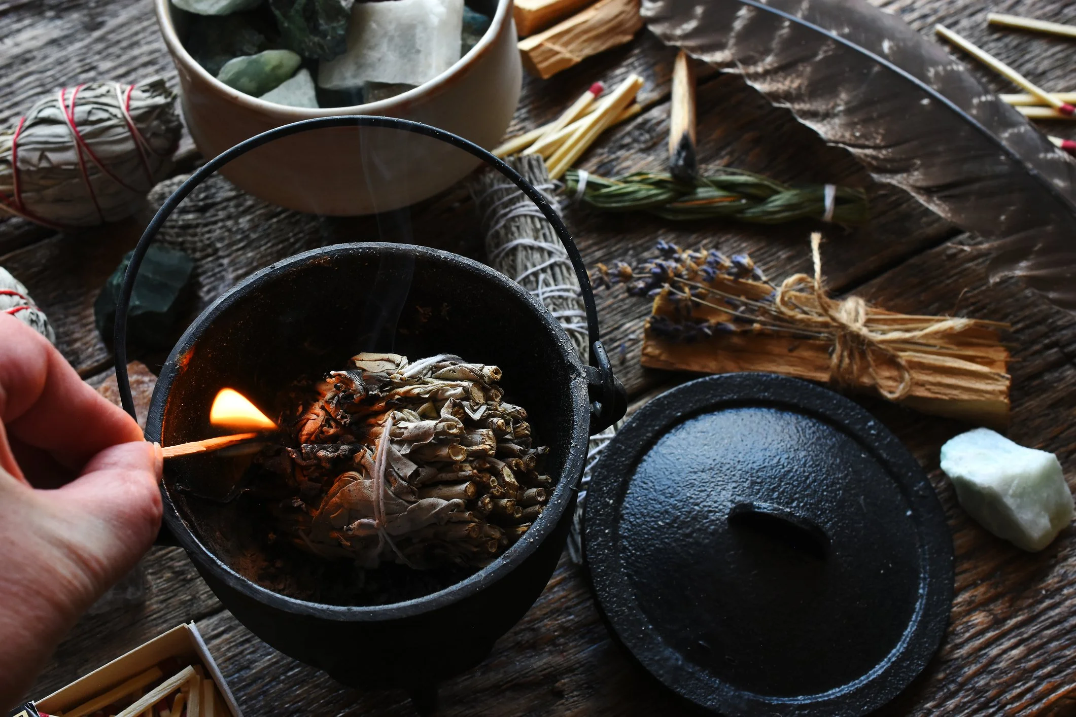 A person lighting a bundle of sage with a match in a black cauldron, surrounded by ritual items such as dried herbs, a feather, and stones on a wooden surface.