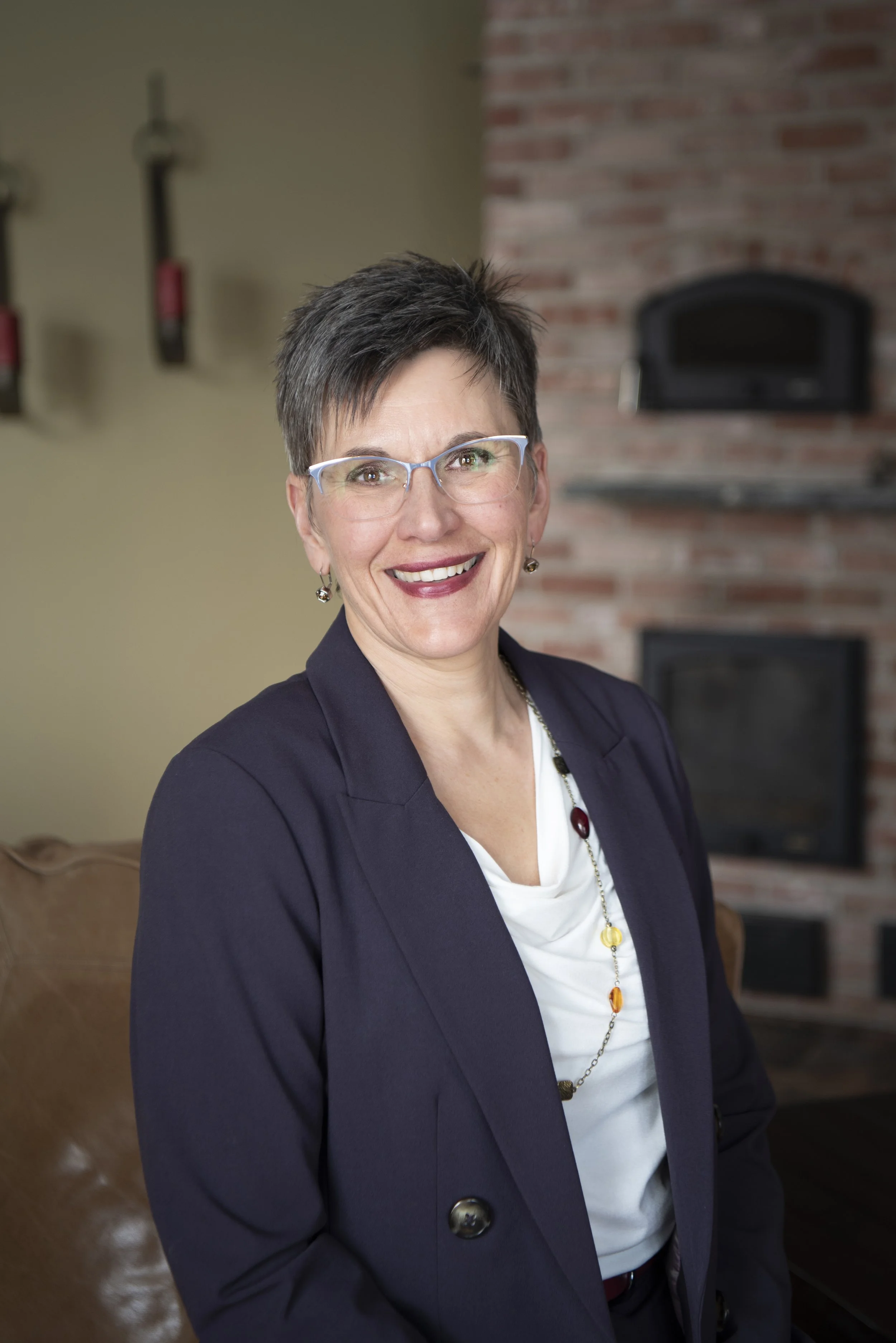 A smiling woman with short dark hair and glasses, wearing a navy blazer and a white blouse, in a room with a brick fireplace in the background.