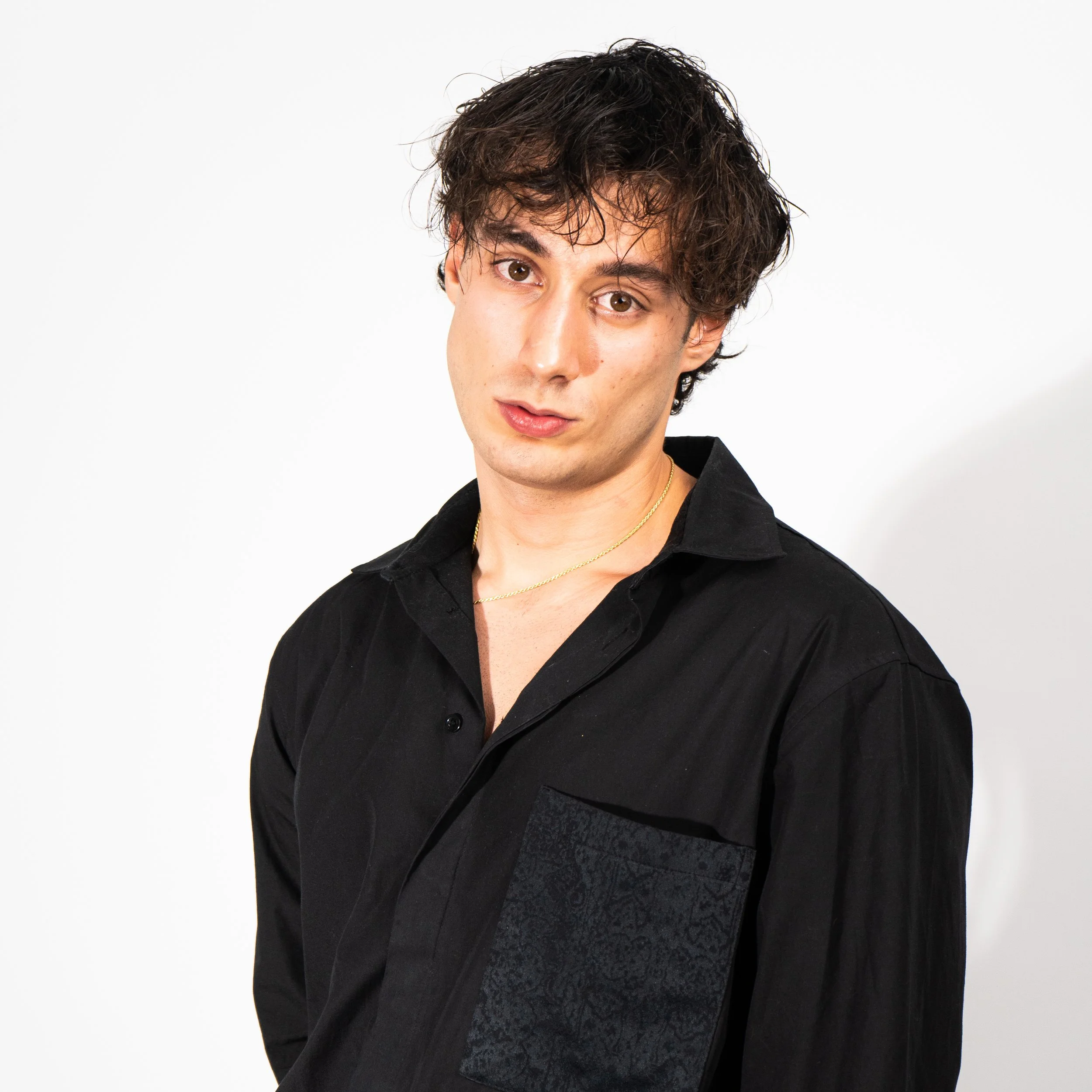 Young man with messy dark hair wearing a black shirt with a patterned pocket and a gold chain, posing against a plain white background.