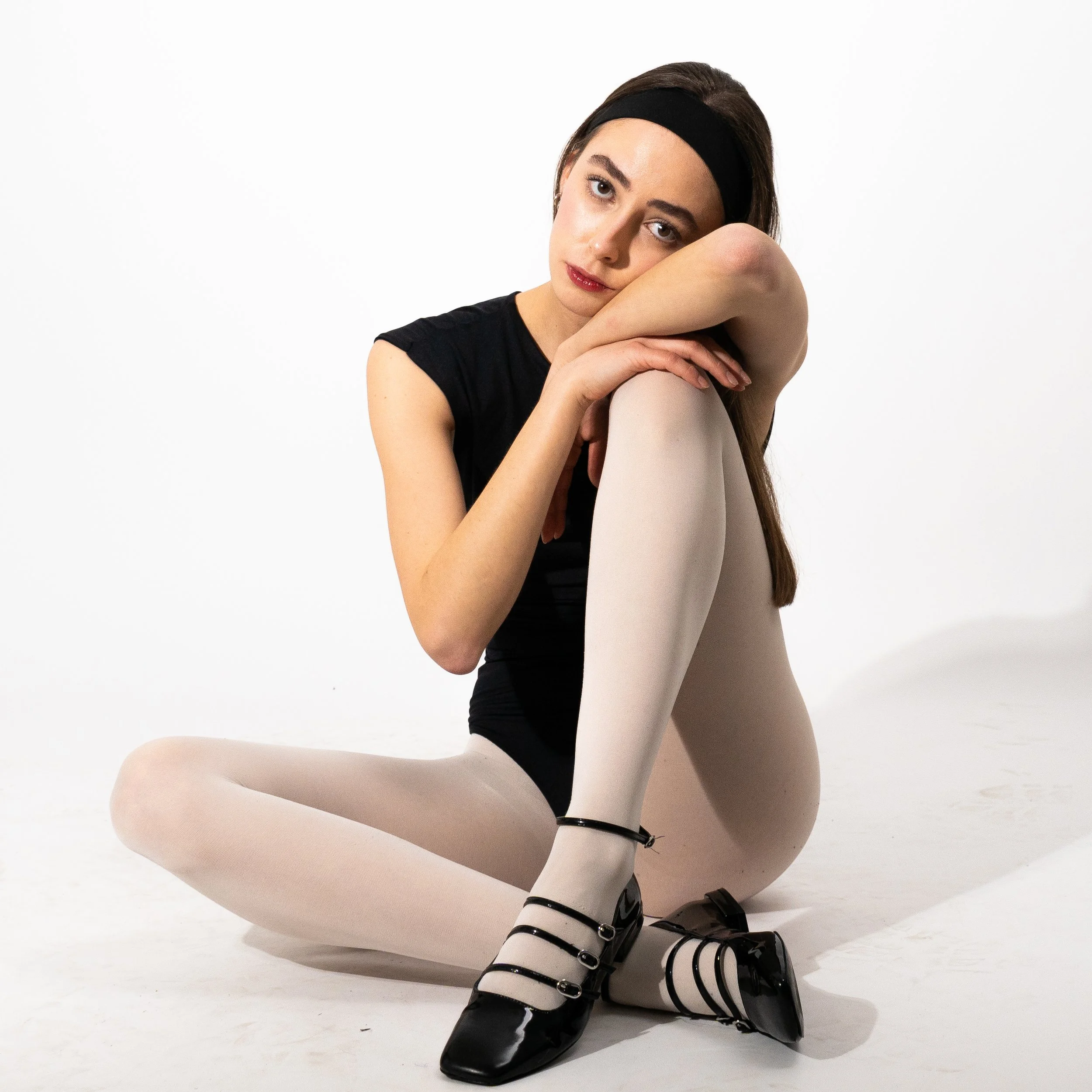 A young woman with long brown hair, wearing a black headband, black sleeveless top, beige tights, and black shoes, sitting on the floor against a white background, resting her head on her knees and looking at the camera.