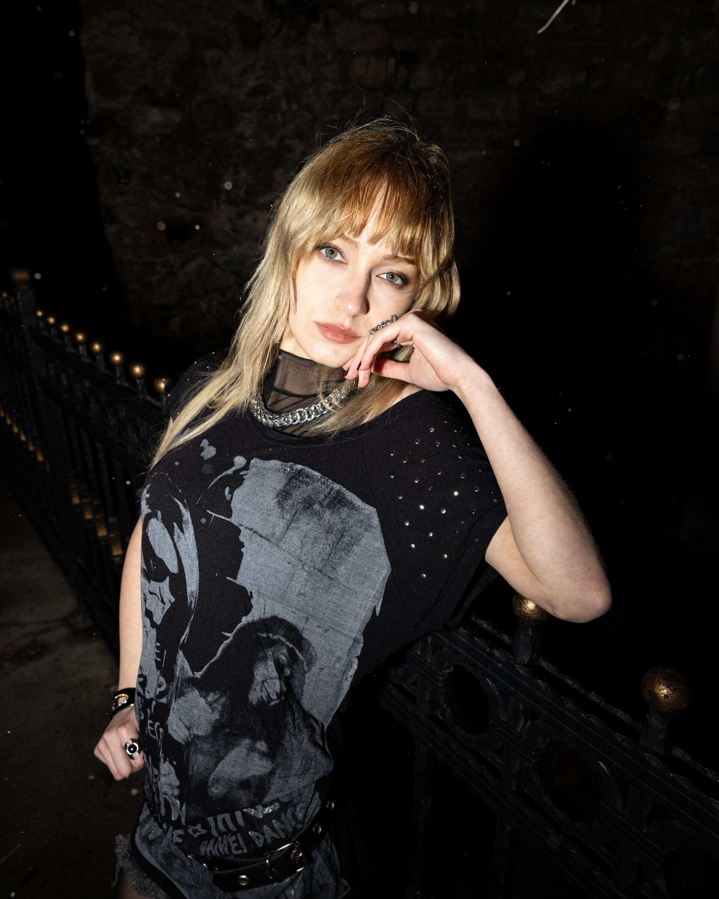 A young woman with blonde hair standing next to a black iron fence. She is wearing a black graphic t-shirt and accessories, including a silver chain necklace and rings. She has a confident expression and is resting her face on her hand.