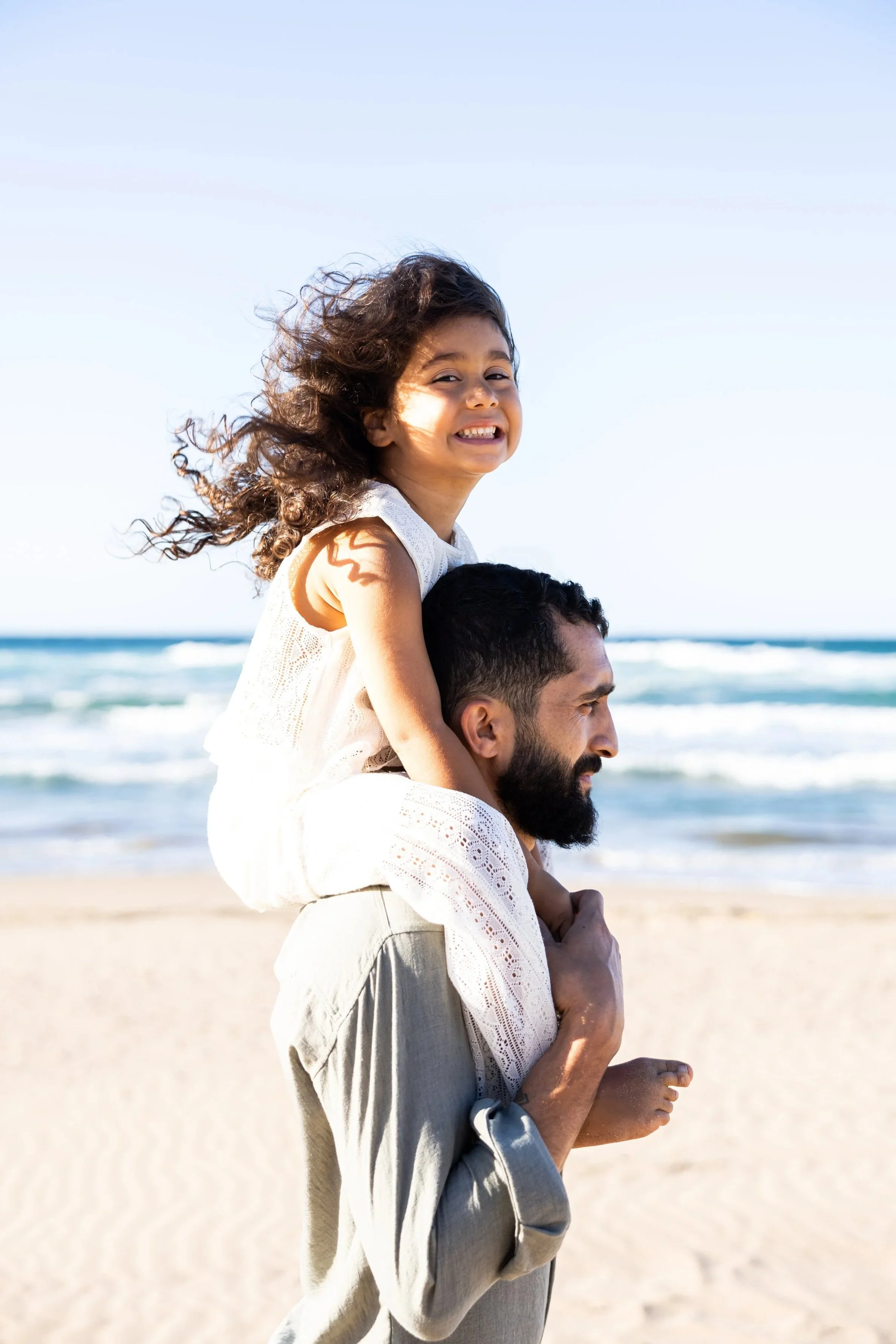 A man carrying a young girl on his shoulders at the beach, with ocean waves in the background.