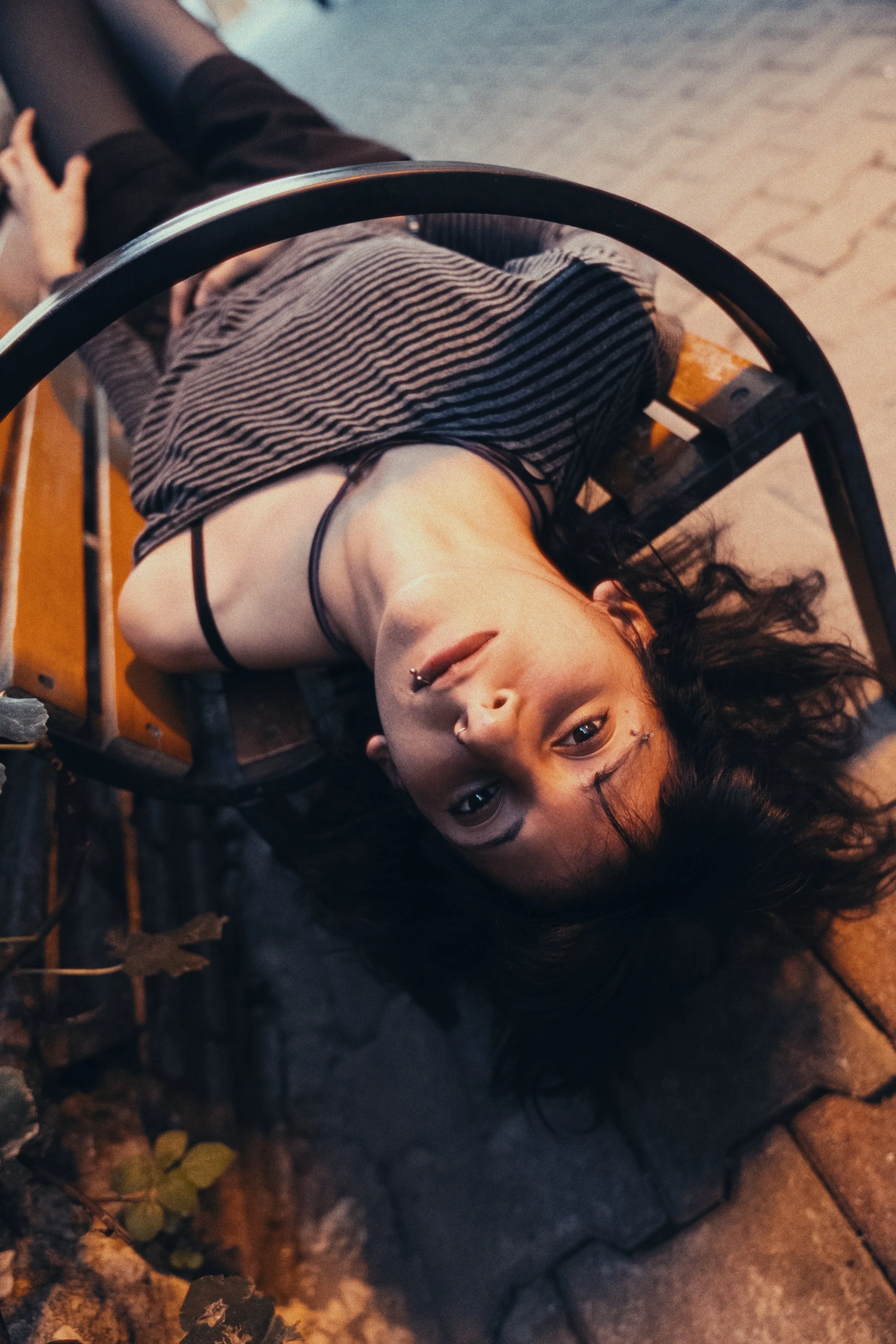 A woman with dark curly hair and a nose piercing lying upside down on a park bench, looking at the camera, at night.