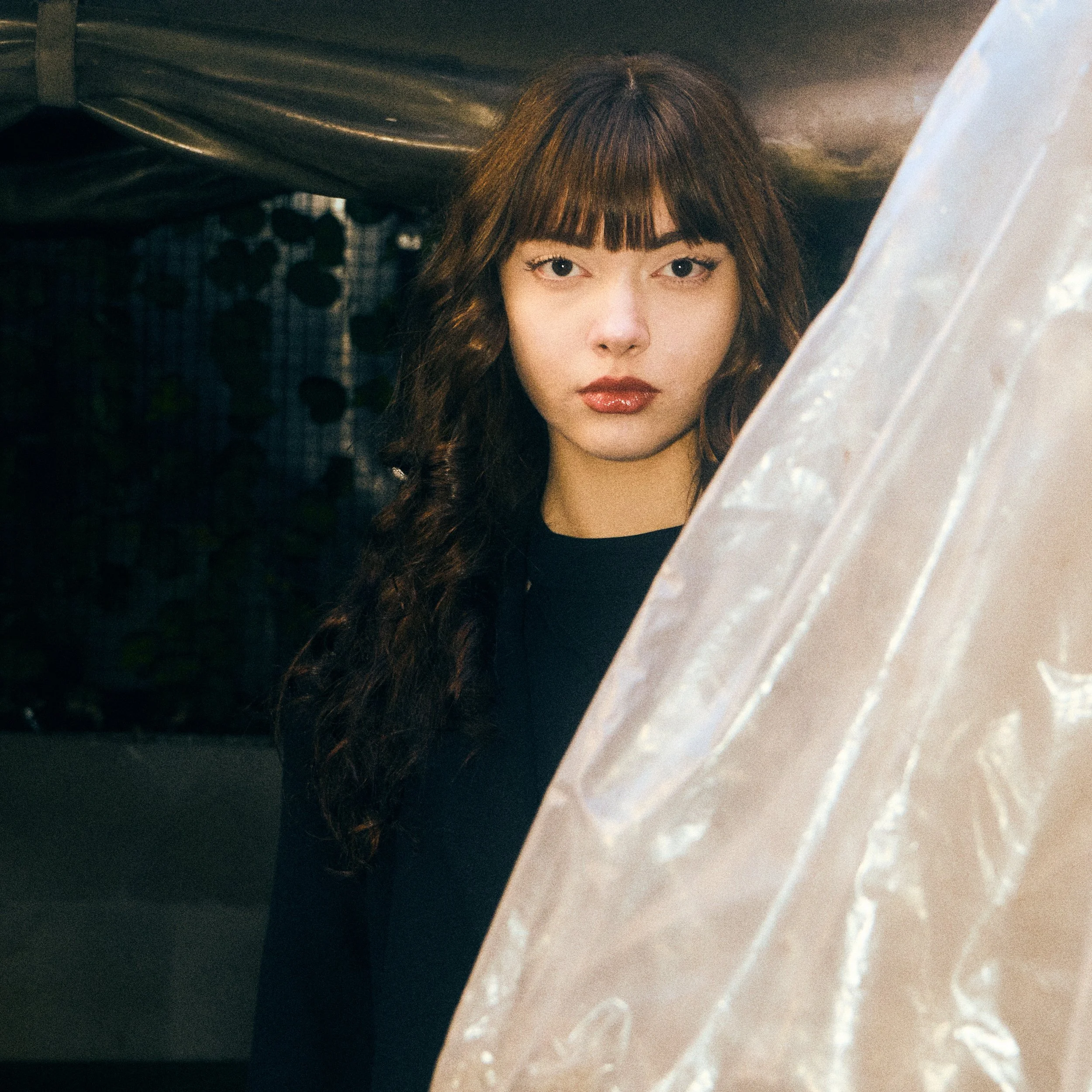 A woman with long, wavy dark hair and bangs, wearing a black top, looking directly at the camera in front of a dark background with a plastic sheet partially covering her face.