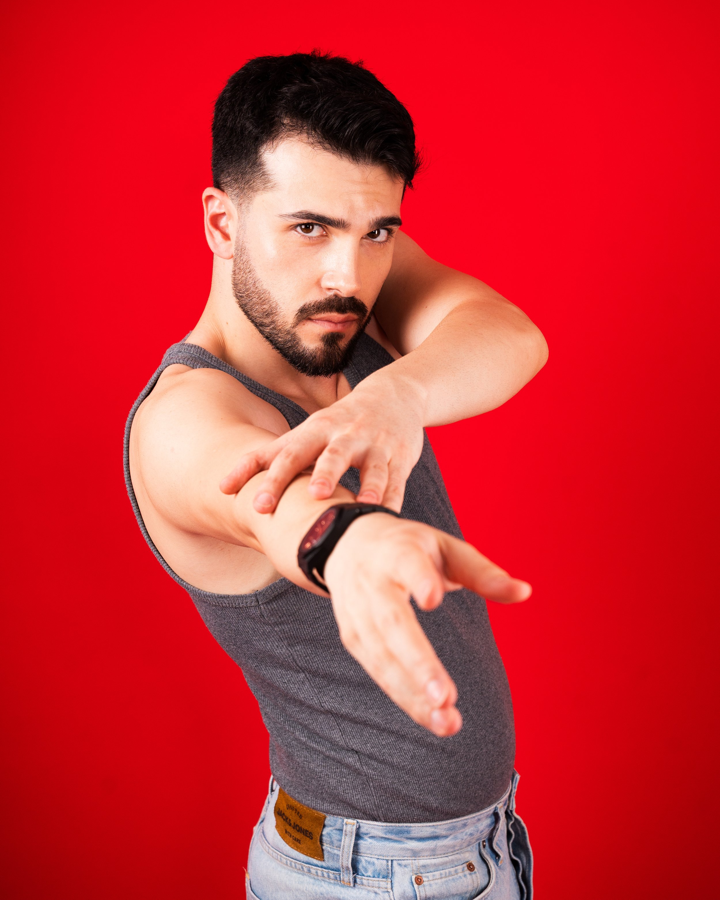 A man with dark hair and a beard, wearing a gray tank top, is stretching his arm with a serious expression on a red background.