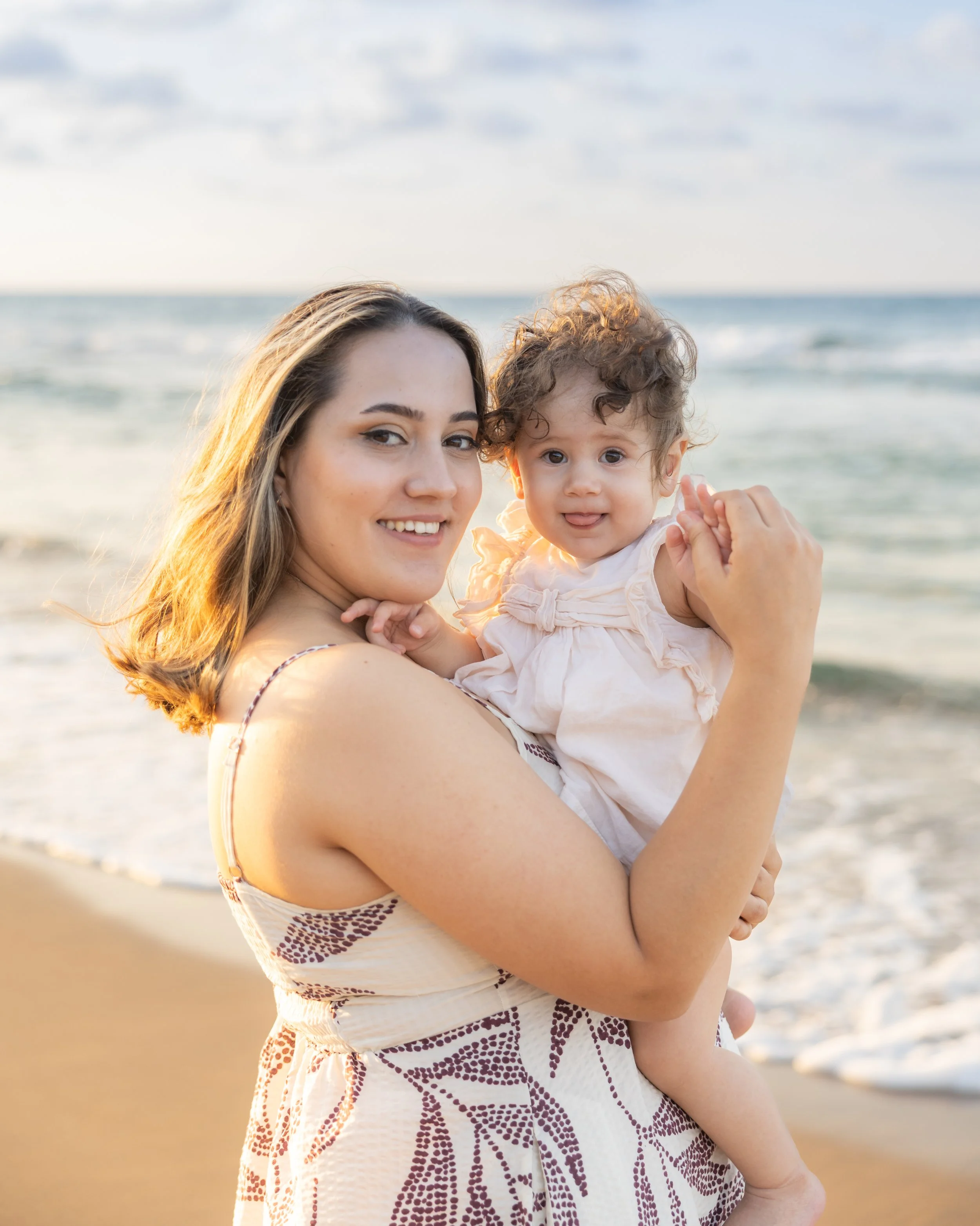 A woman holding a toddler girl on a beach at sunset, with the ocean in the background.