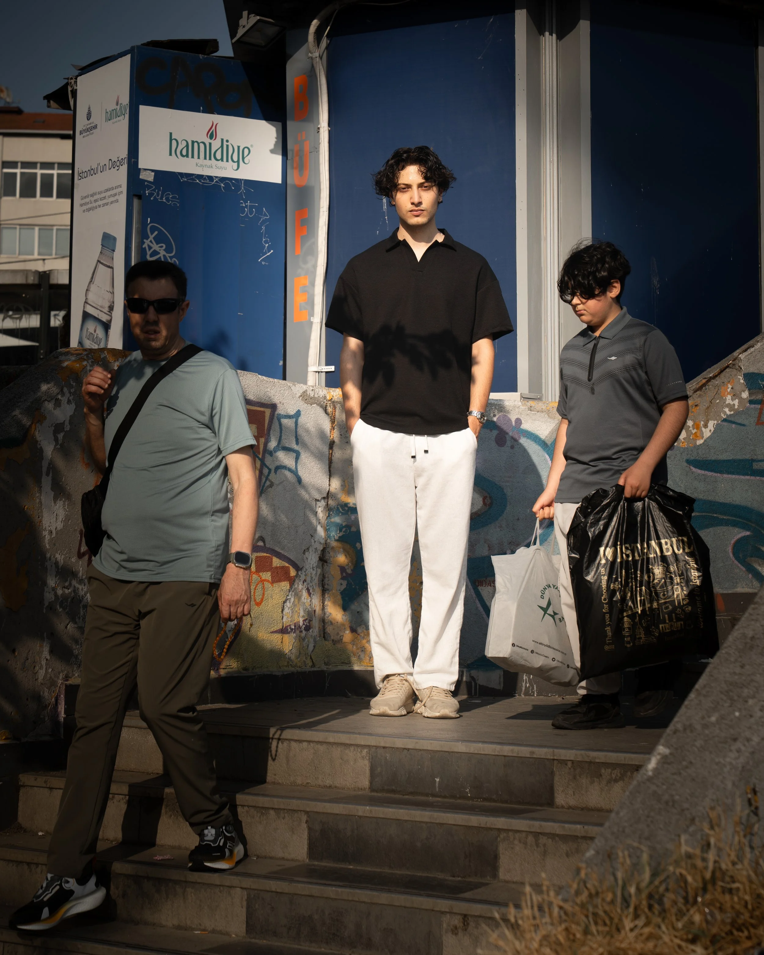 Three young men standing on stairs outside near blue storefronts, with one man in the foreground walking down stairs and two others standing behind him, one holding shopping bags, in sunlight.