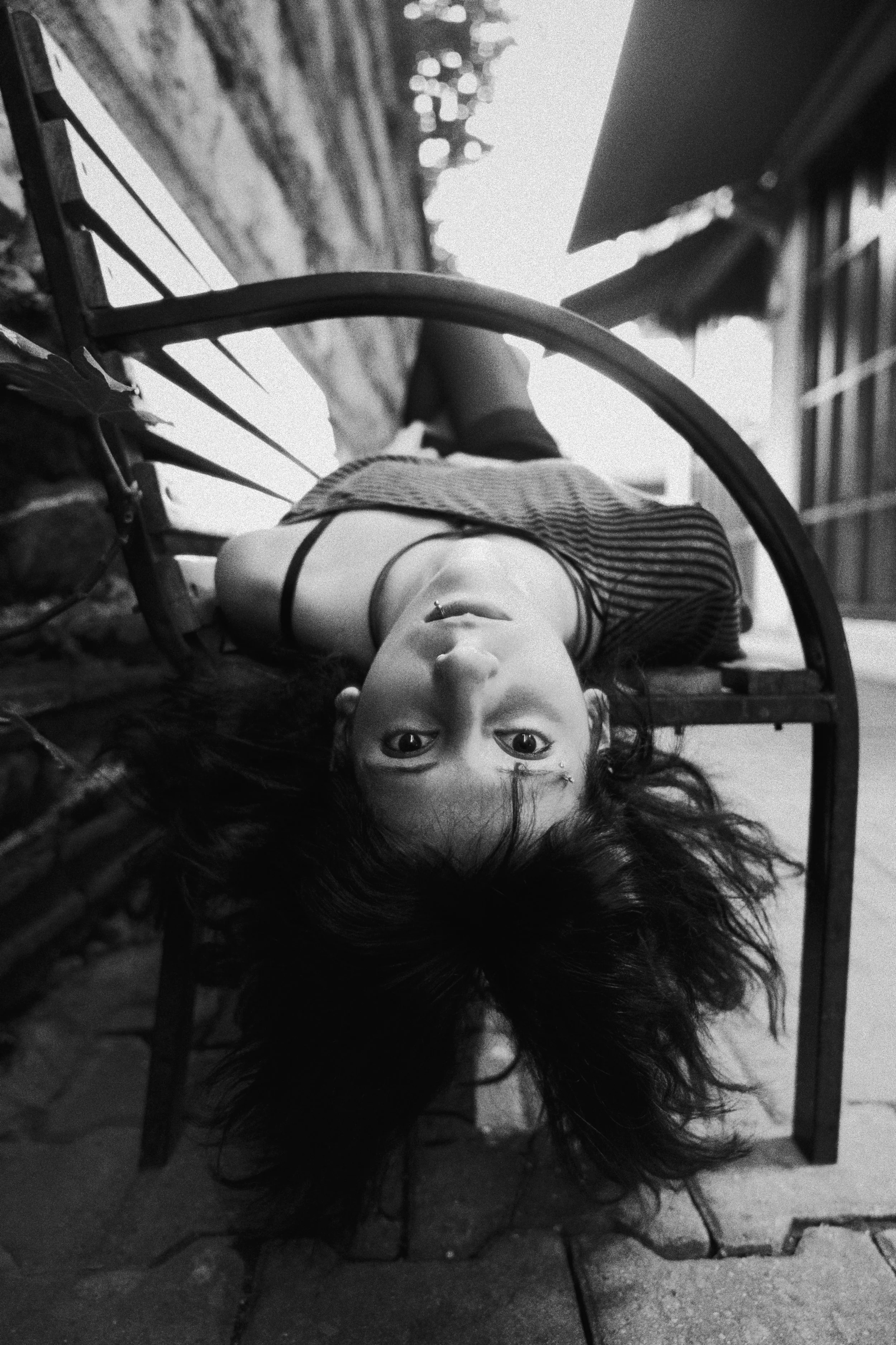 Black and white photo of a woman lying upside down on a bench with her head hanging off the edge, looking directly at the camera.