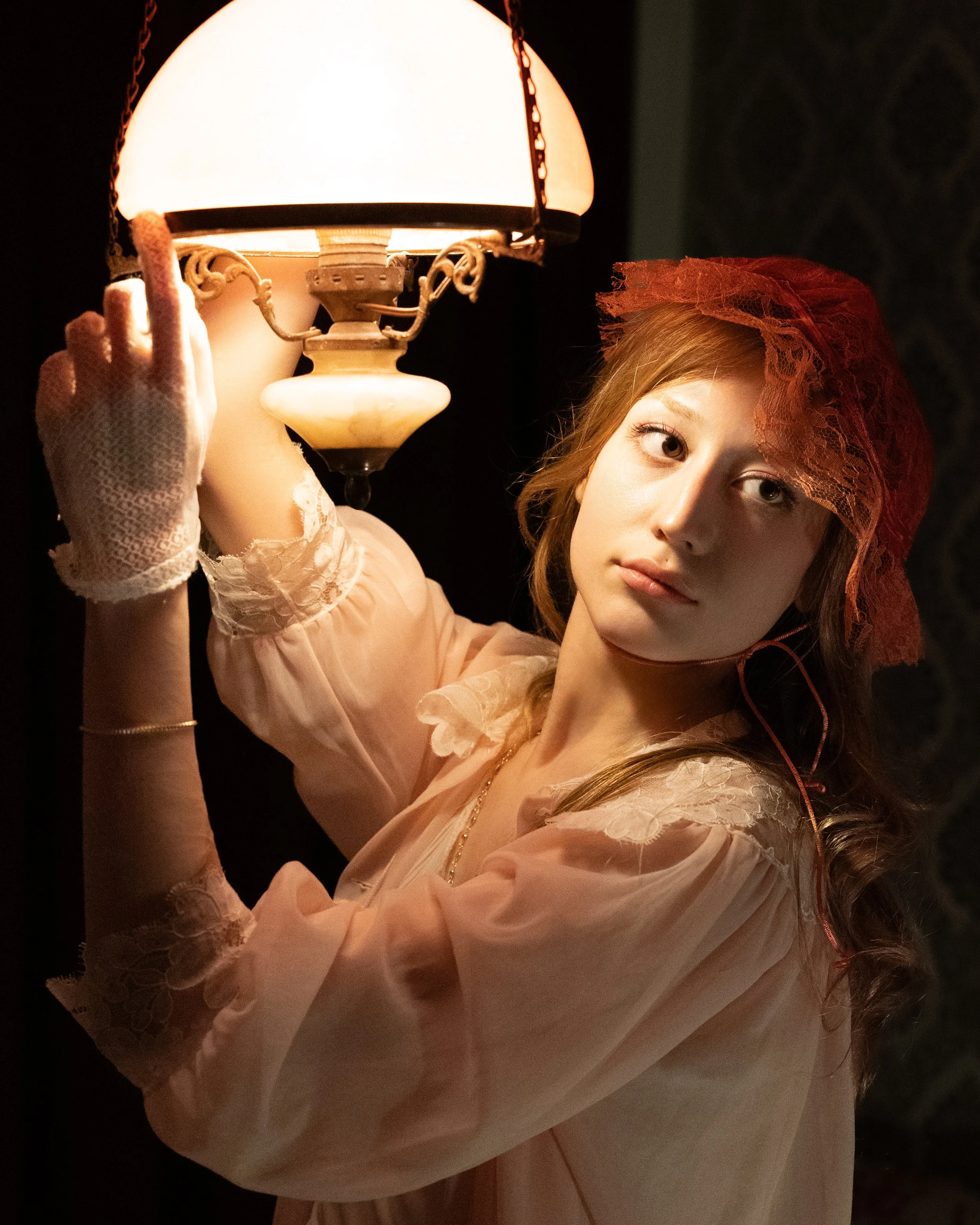 Young woman in vintage clothing with lace gloves and red lace head covering, holding an ornate table lamp with a warm glow.