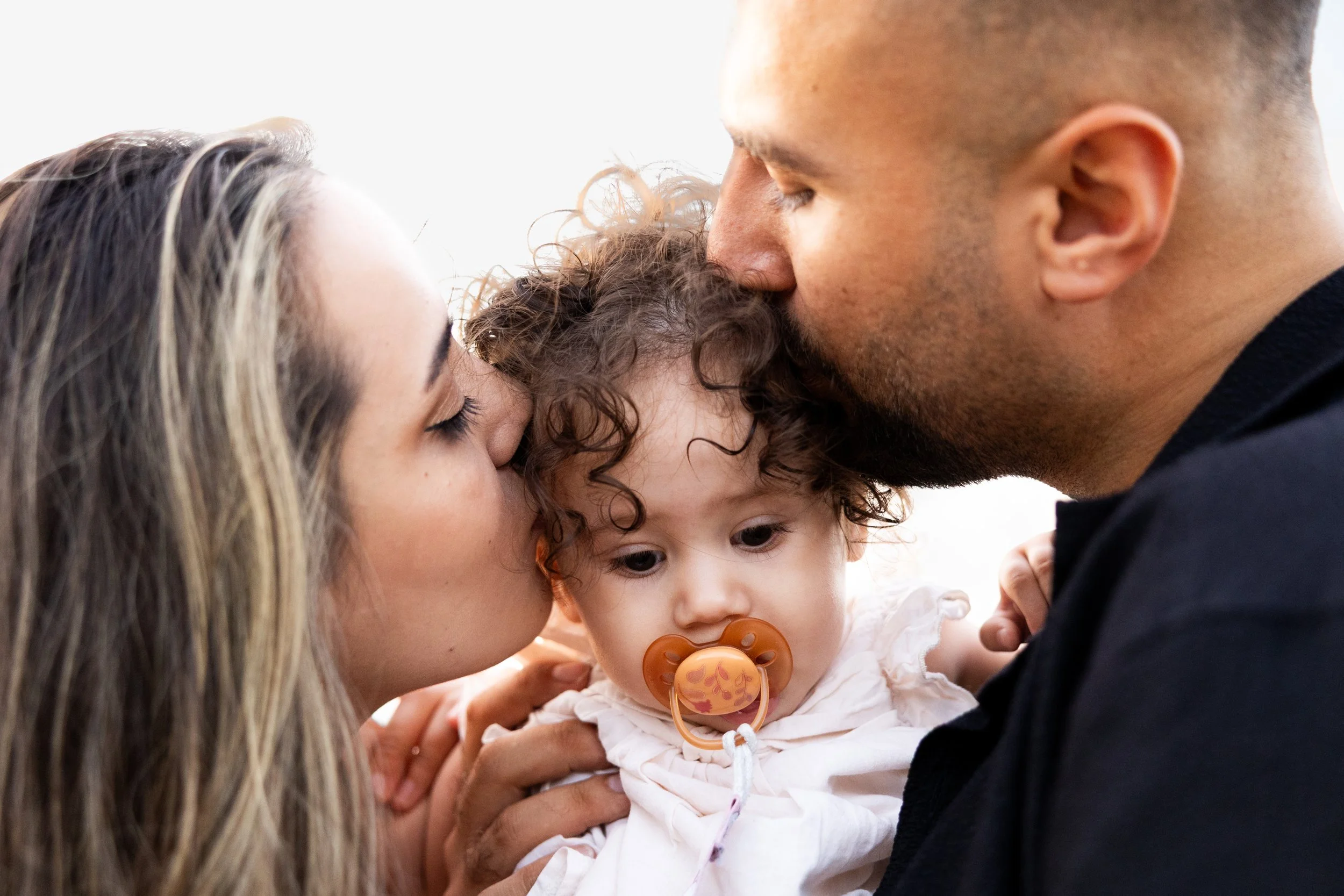 A woman and a man kiss a young girl with curly hair and a pacifier, holding her gently.