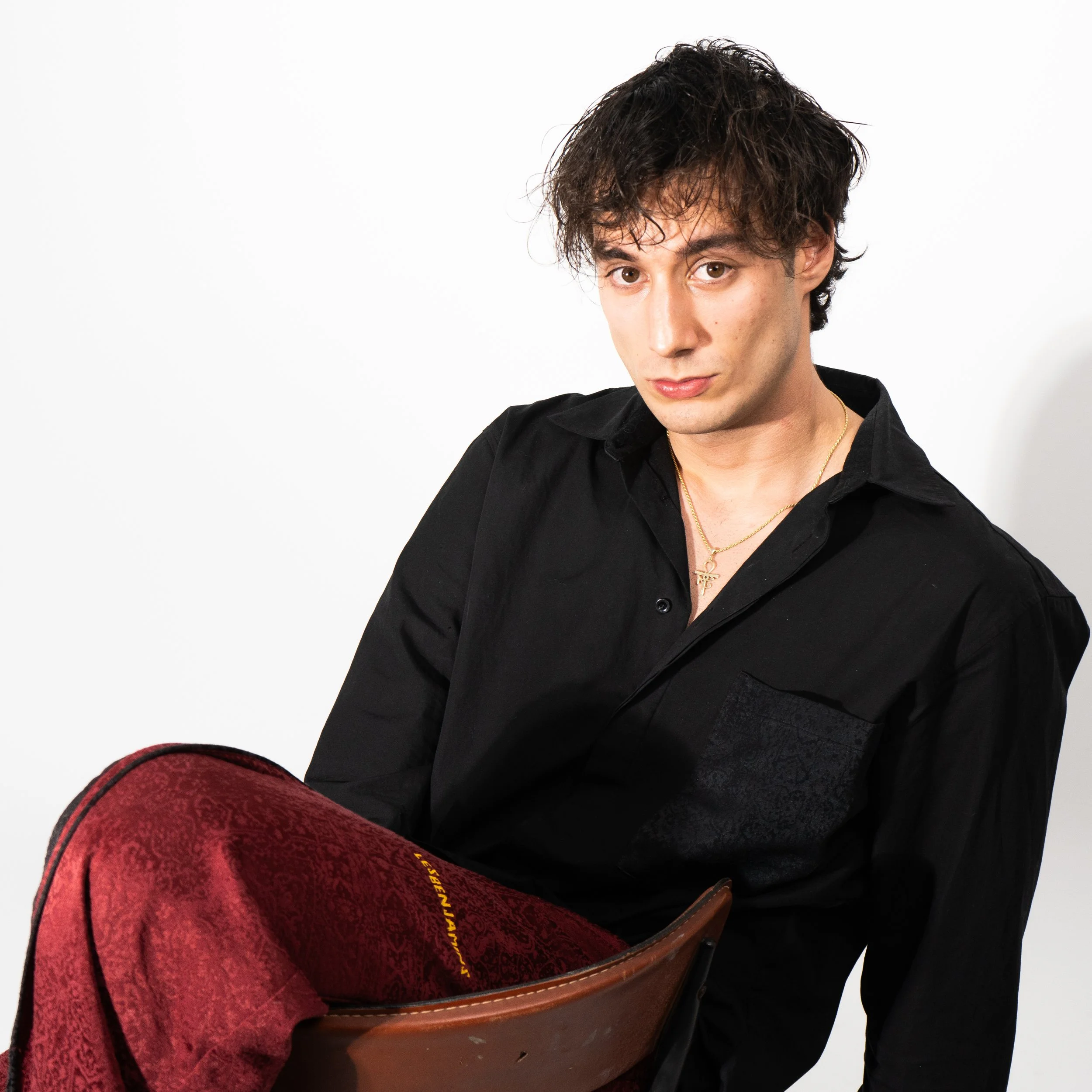 Young man with dark, curly hair wearing a black shirt and gold necklace, sitting on a chair against a white background, holding a red patterned blanket.