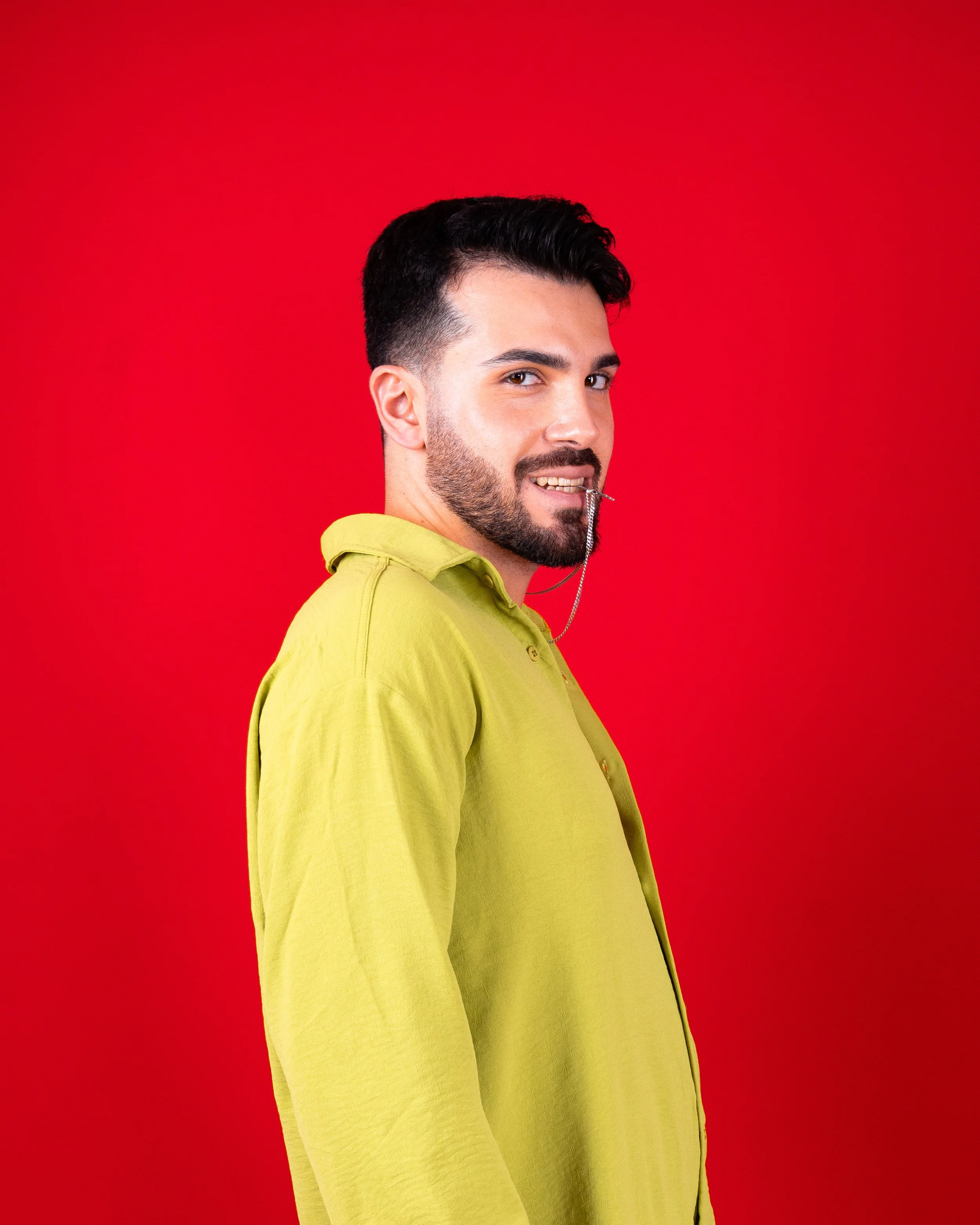 A man with dark hair and a beard wearing a yellow shirt, smiling with a chain in his mouth, against a red background.