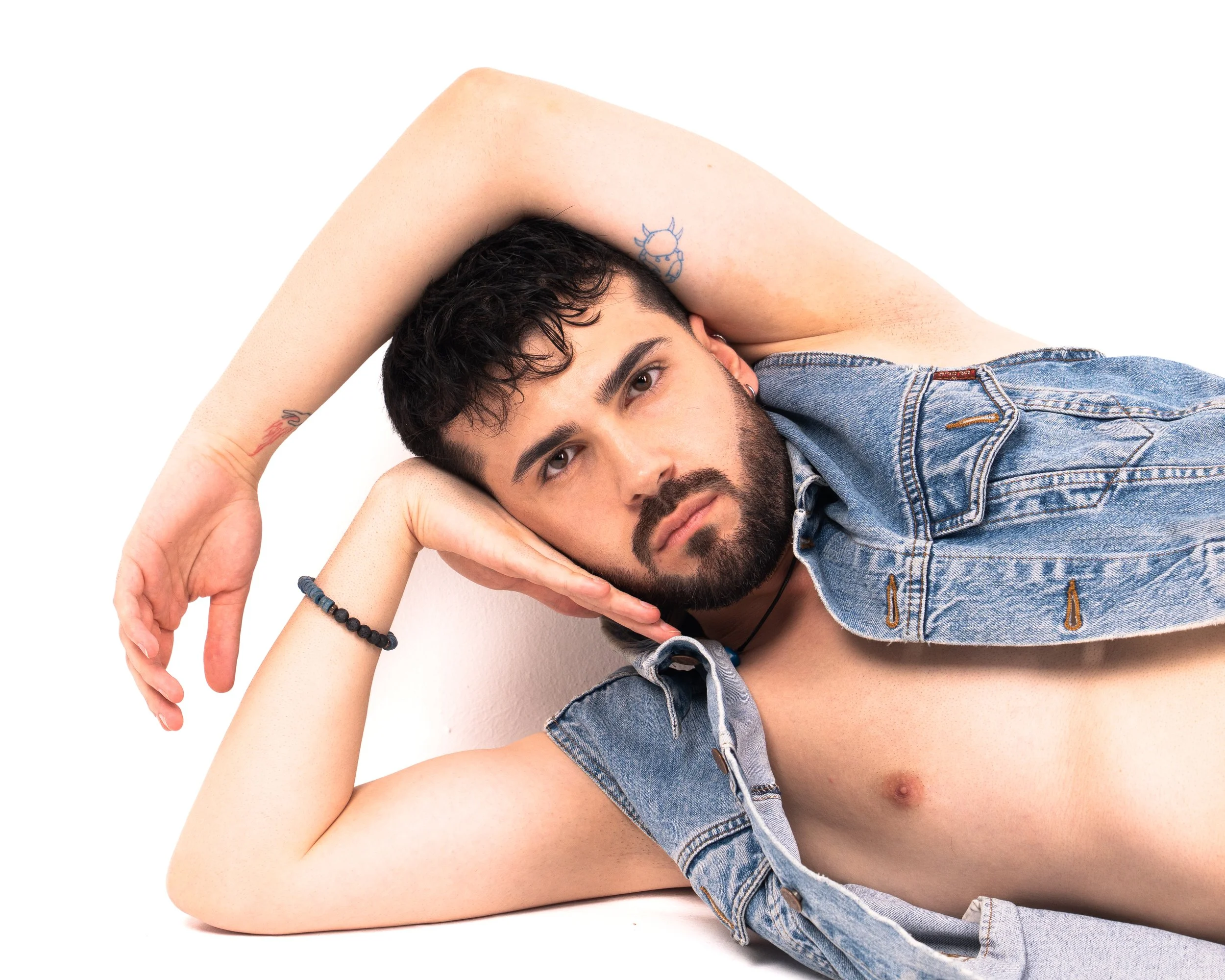 A man with dark curly hair and a beard, lying on his side with his head propped up by his hand, wearing an open denim vest, against a white background.