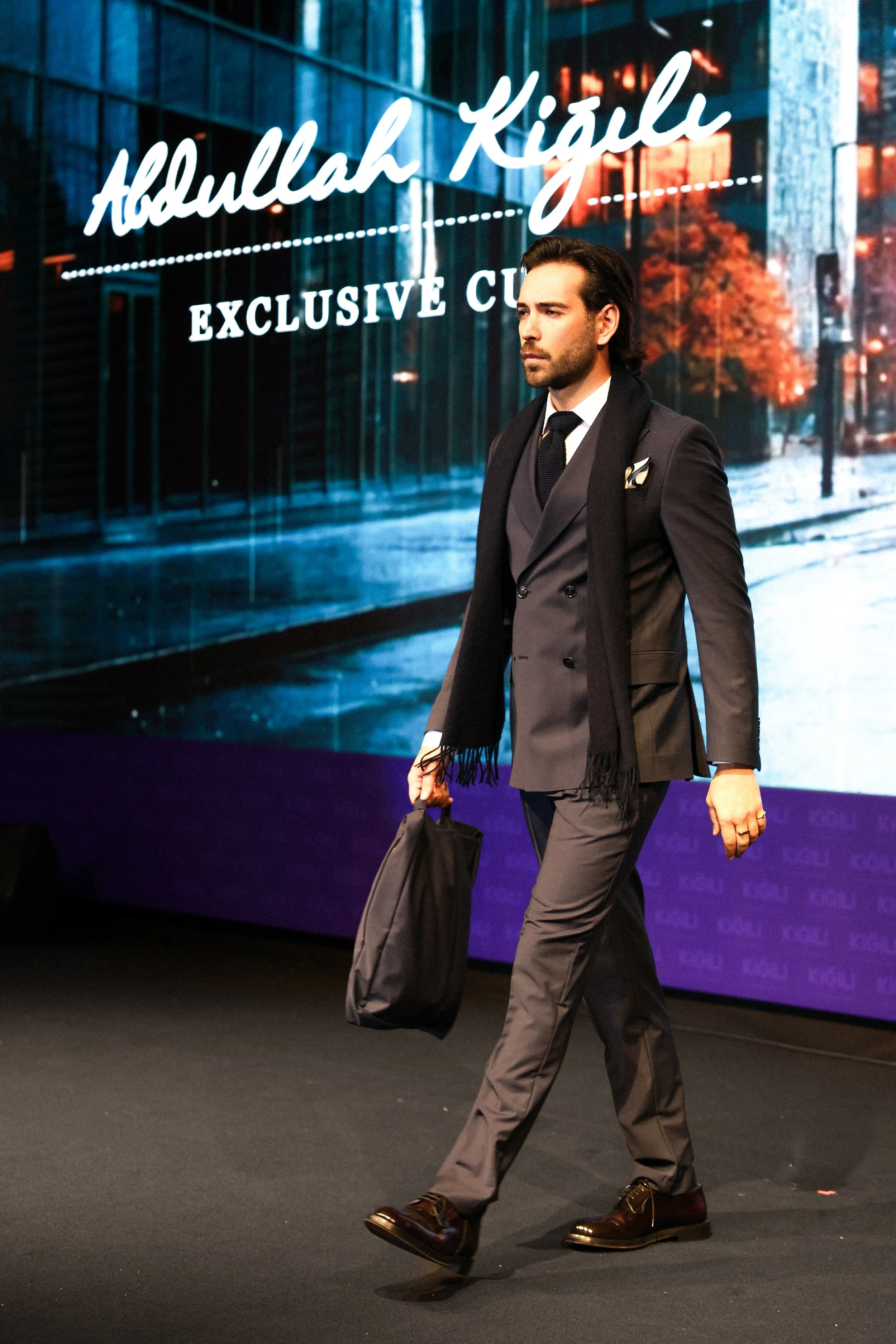 A man in a gray business suit walking on stage, carrying a black bag, with a large digital display behind him showing the text 'Abdullah Kızıl' and 'Exclusive Club'.
