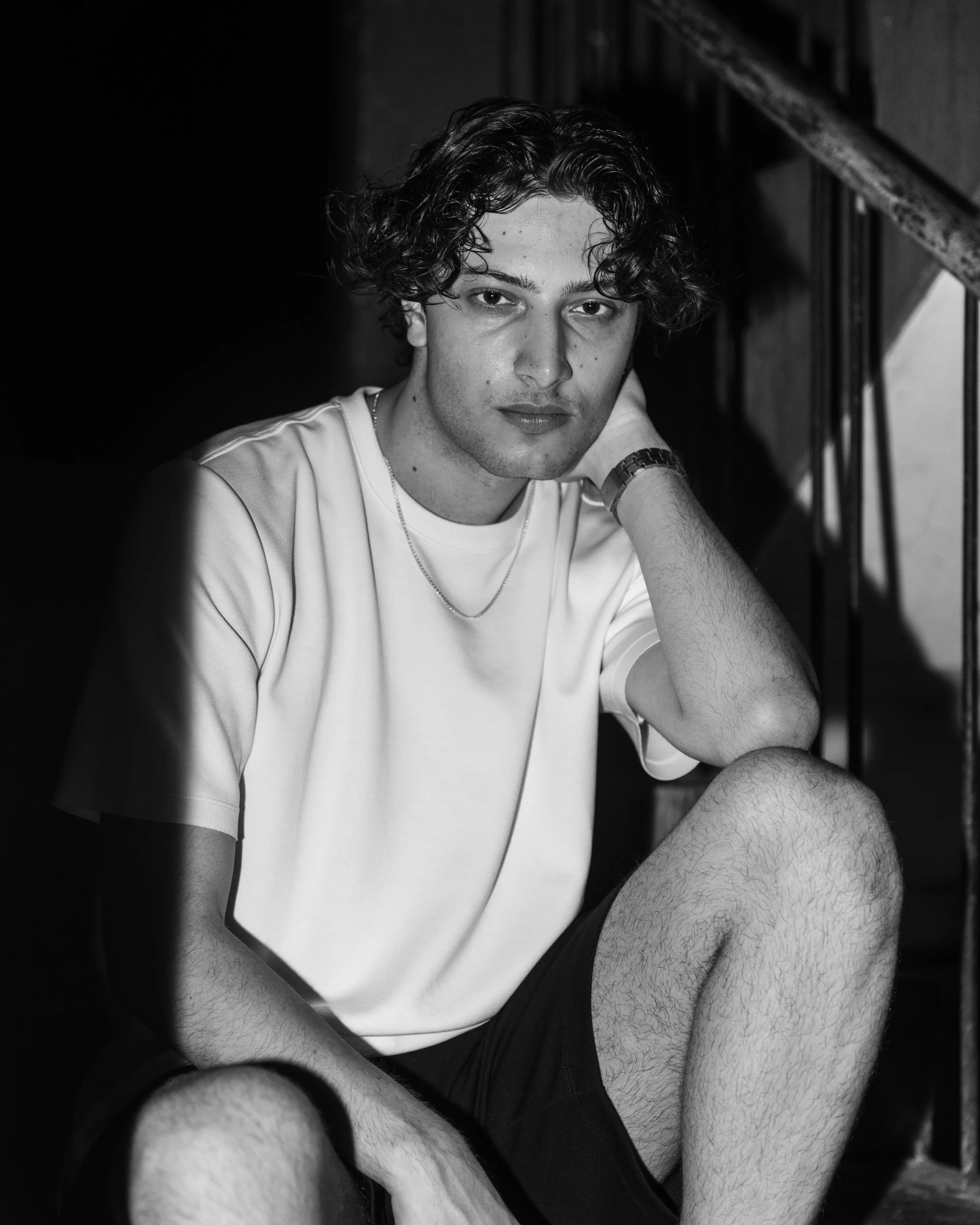 Black and white photo of a young man with curly hair, sitting on stairs, wearing a white t-shirt, shorts, and a watch, with a contemplative expression.
