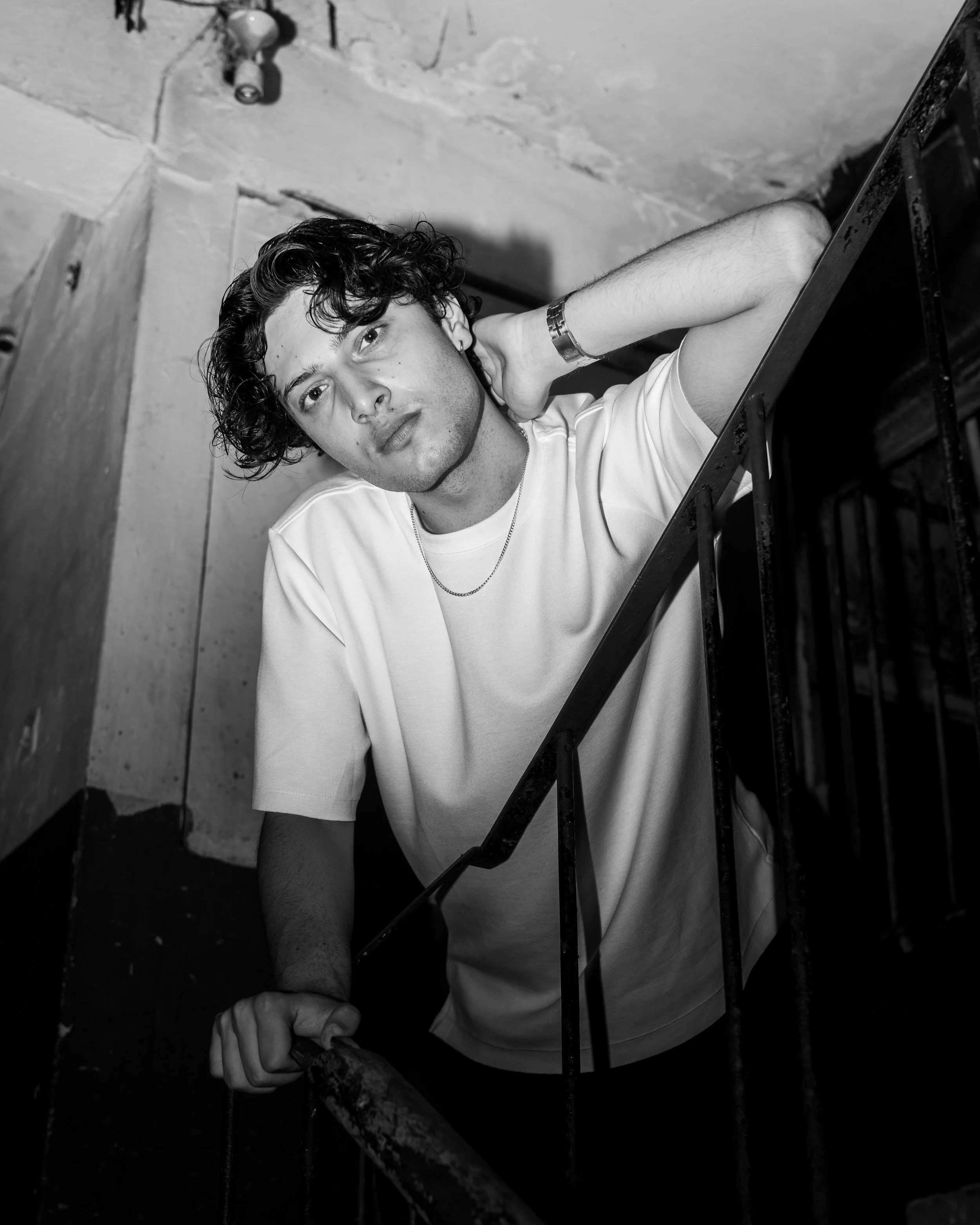 A young man with curly hair, wearing a white t-shirt, leaning on a staircase railing, looking directly at the camera with a relaxed expression, in a black and white photo.