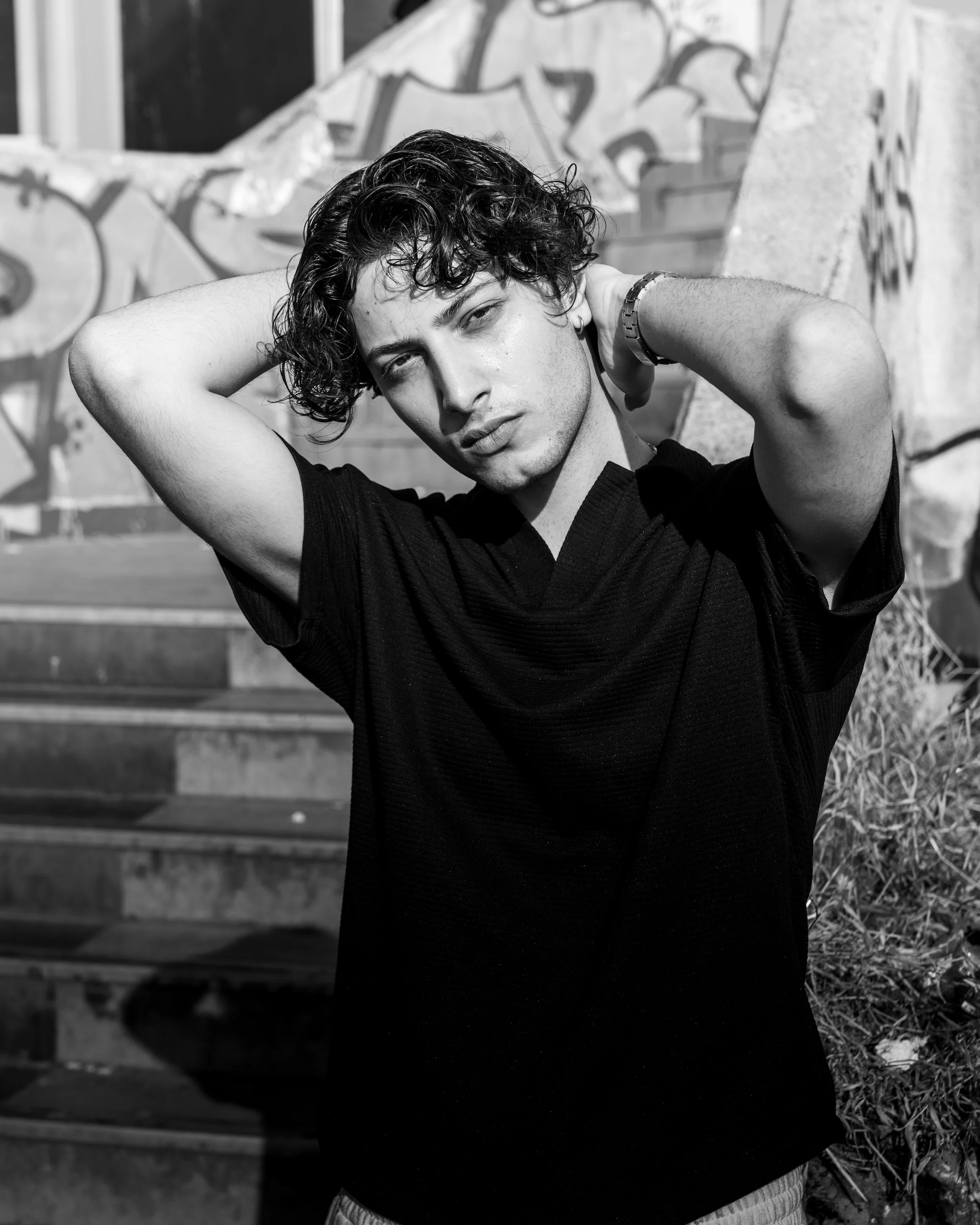 A young man with curly hair posing outdoors, with graffiti on a wall and stairs in the background.