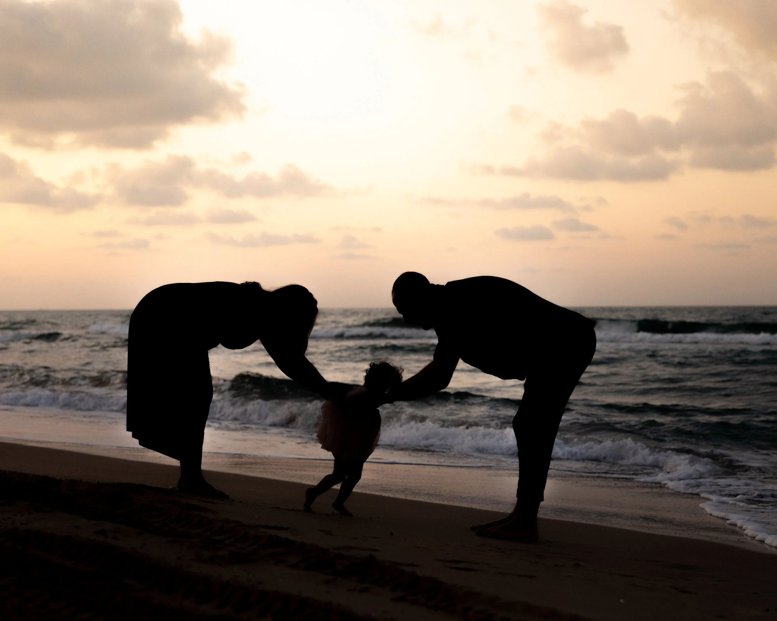 Silhouettes of two adults and a child holding hands on a beach during sunset, with ocean waves in the background.