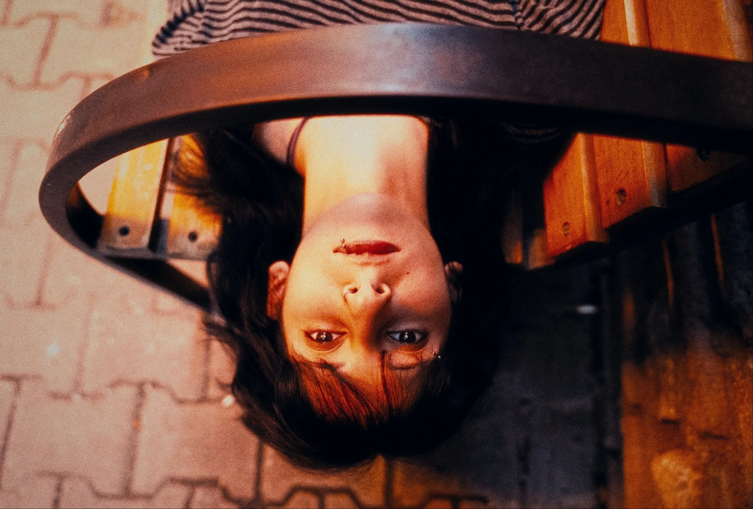 A woman with dark hair and piercings, lying on her back under a metal chair, looking directly at the camera with her head tilted back.
