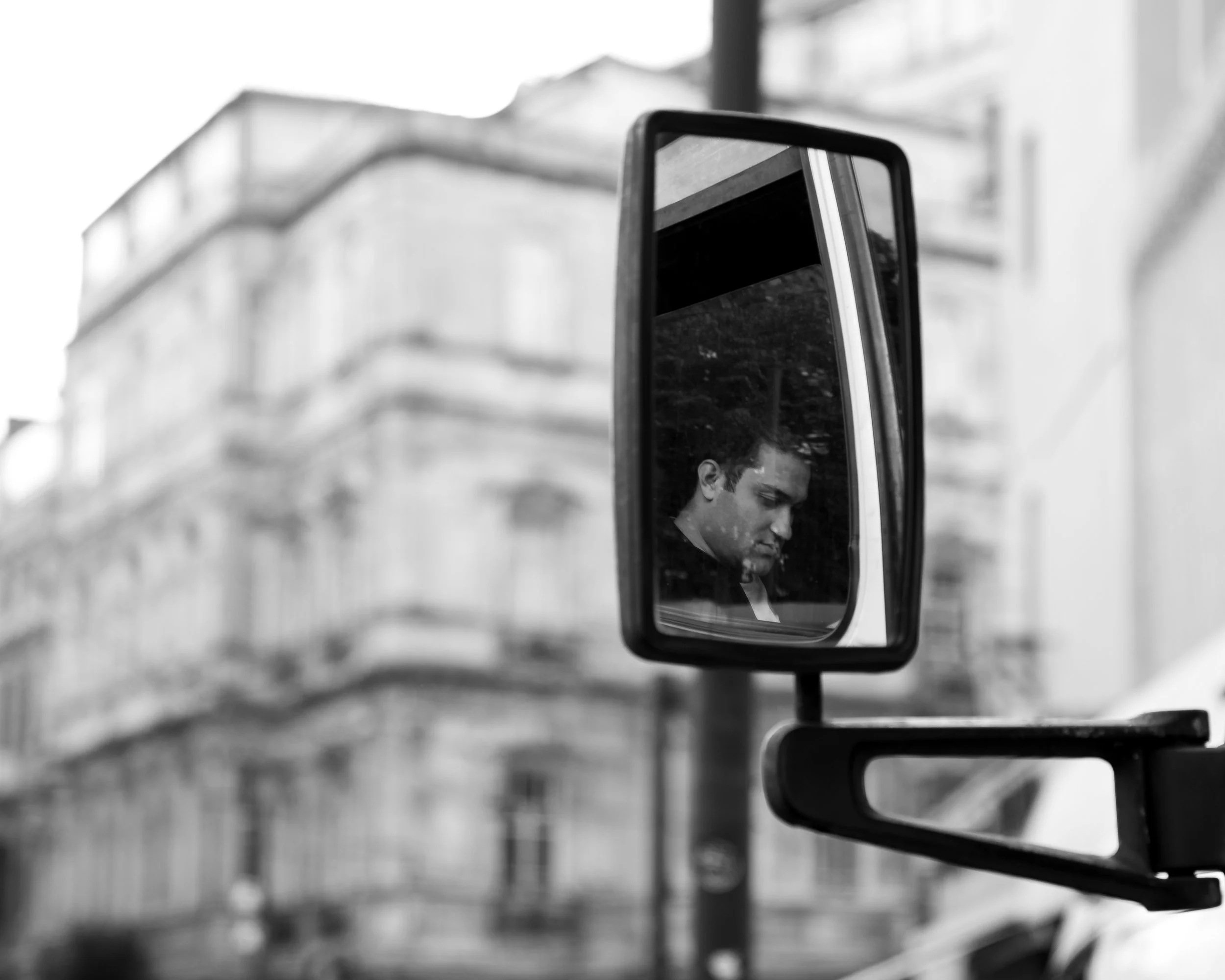 A black-and-white photo of a man’s reflection in a truck's side mirror. The background shows an urban building.