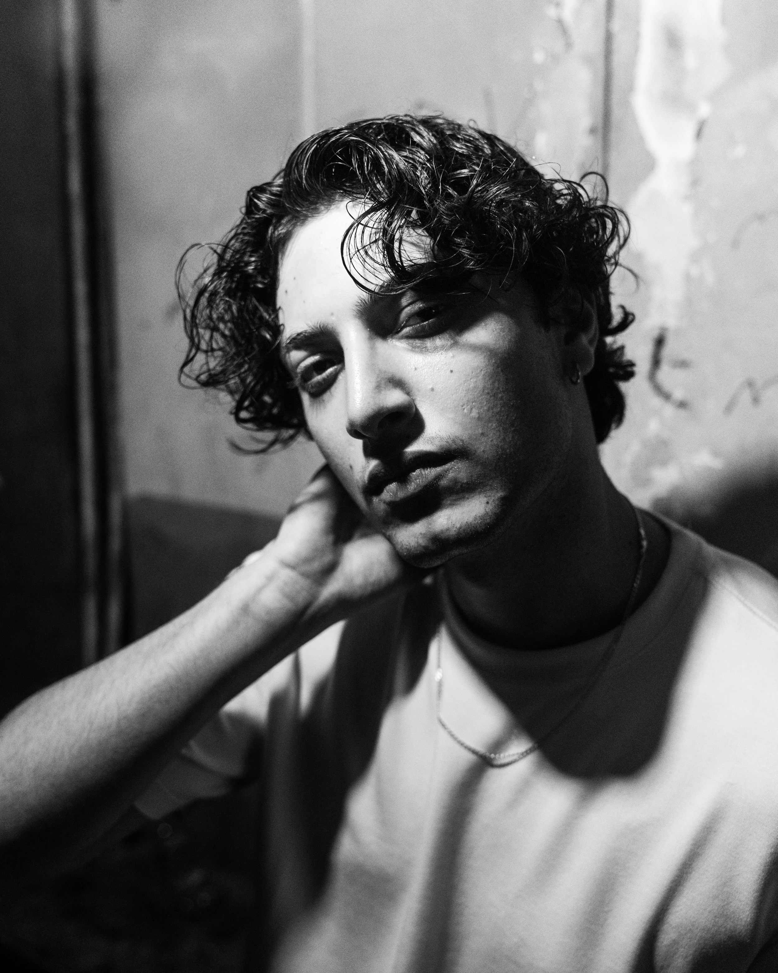 Black and white portrait of a young person with curly hair, looking into the camera, resting their head on their hand, in front of a textured wall.