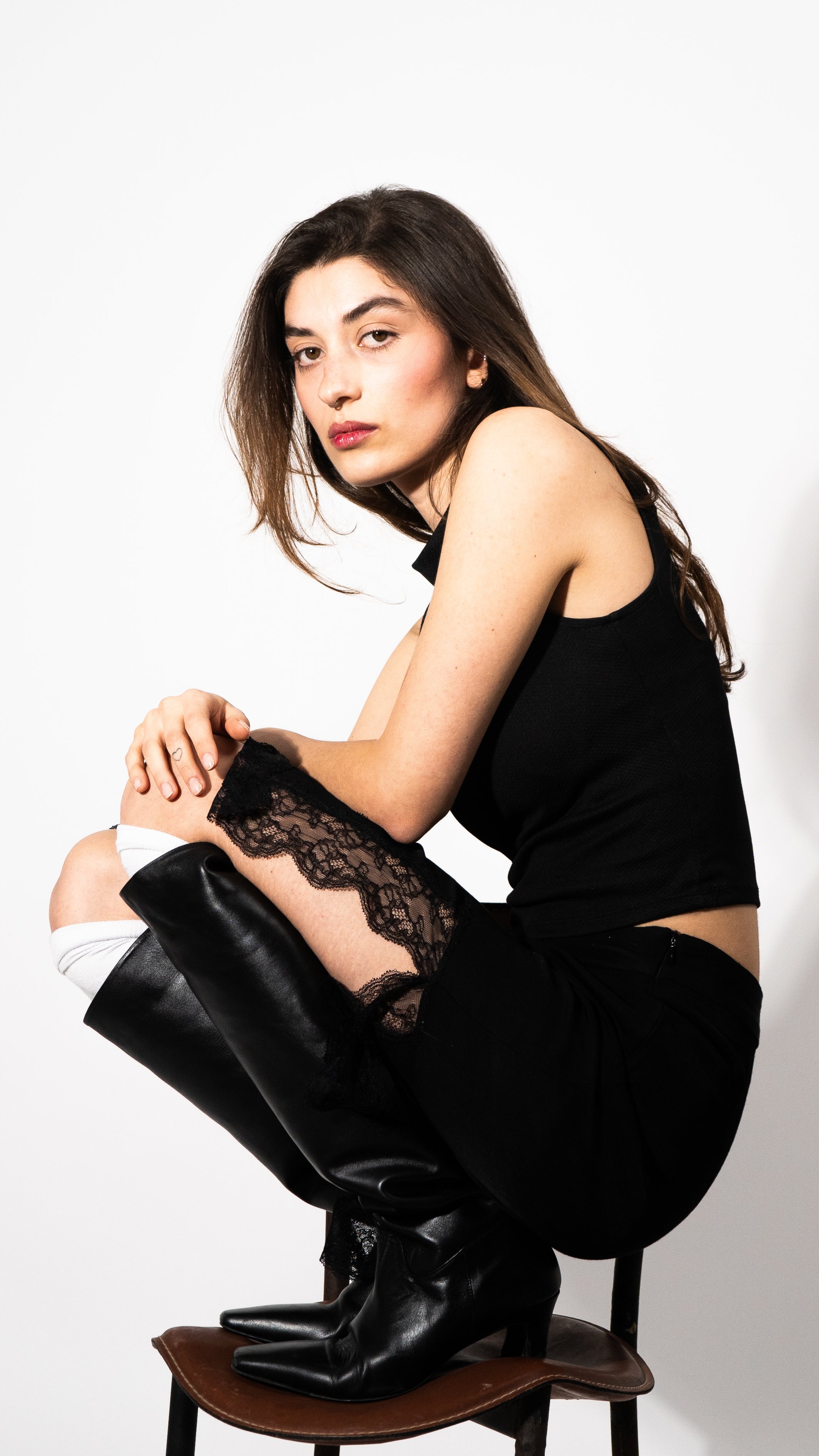 A woman with long brown hair, wearing a black sleeveless top, black pants with lace detail, and black high-heeled boots, sitting on a chair with a plain white background.