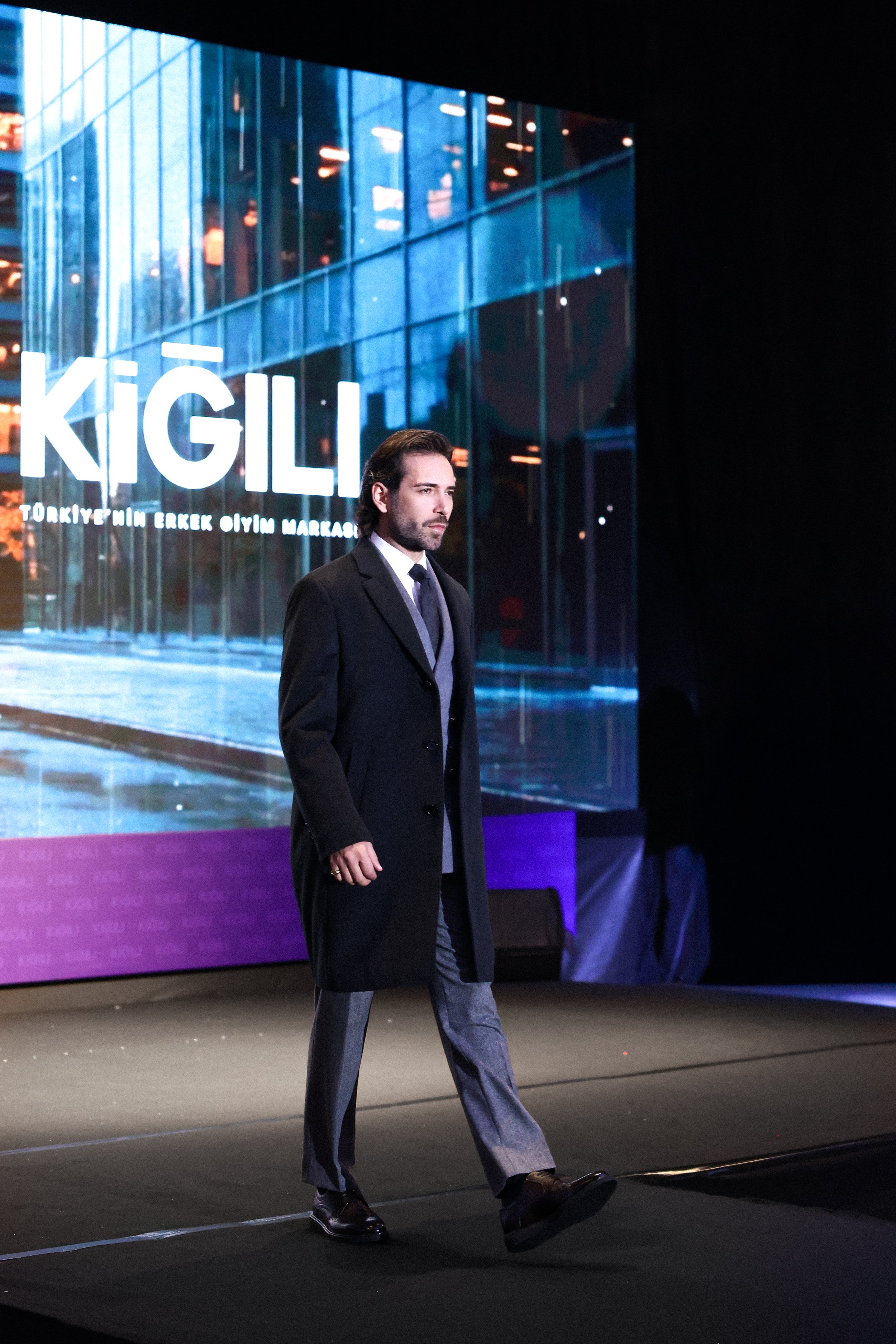 A man with medium-length dark hair and a beard wearing a black coat, gray trousers, and dark shoes walking on a stage in front of a large screen displaying the word 'KİĞILI' with additional Turkish text. The background shows a modern glass building r