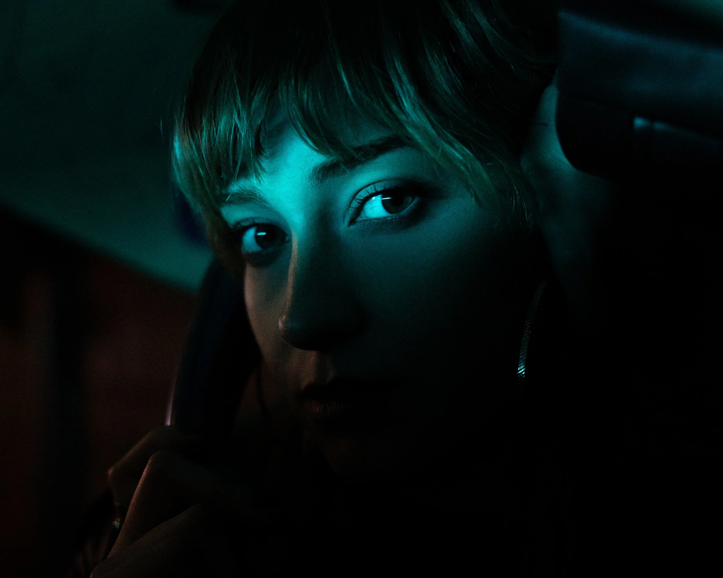 A young woman with short hair illuminated by teal and pink lighting, looking at the camera with a phone held to her ear.