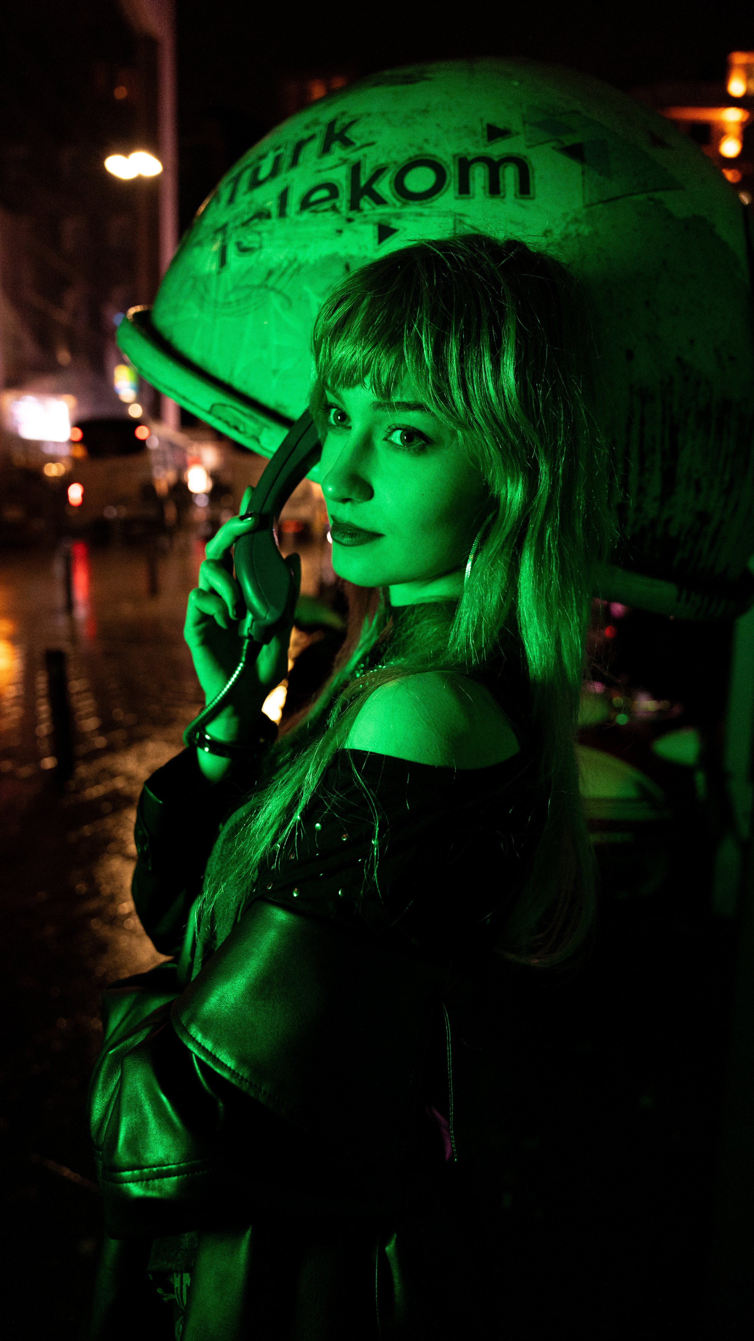 Young woman with long hair holding a green landline phone to her ear, standing under a large hard hat with the logo and text 'PUHIK Telekon,' illuminated with green lighting, in an urban street at night.