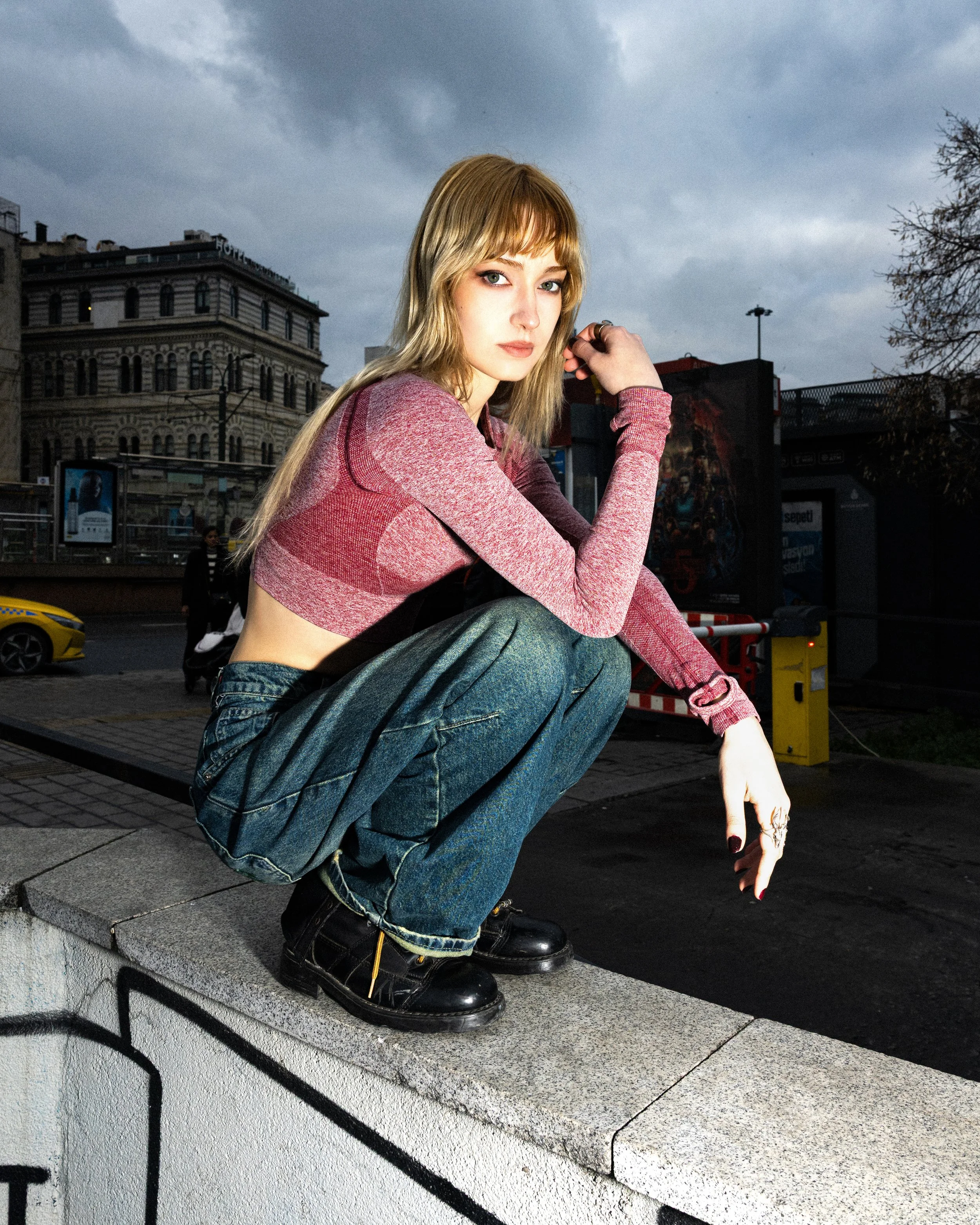 A young woman with blonde hair, wearing a pink long-sleeved top and blue jeans, crouching on a graffiti-wall ledge in an urban setting with cloudy sky and city buildings in the background.