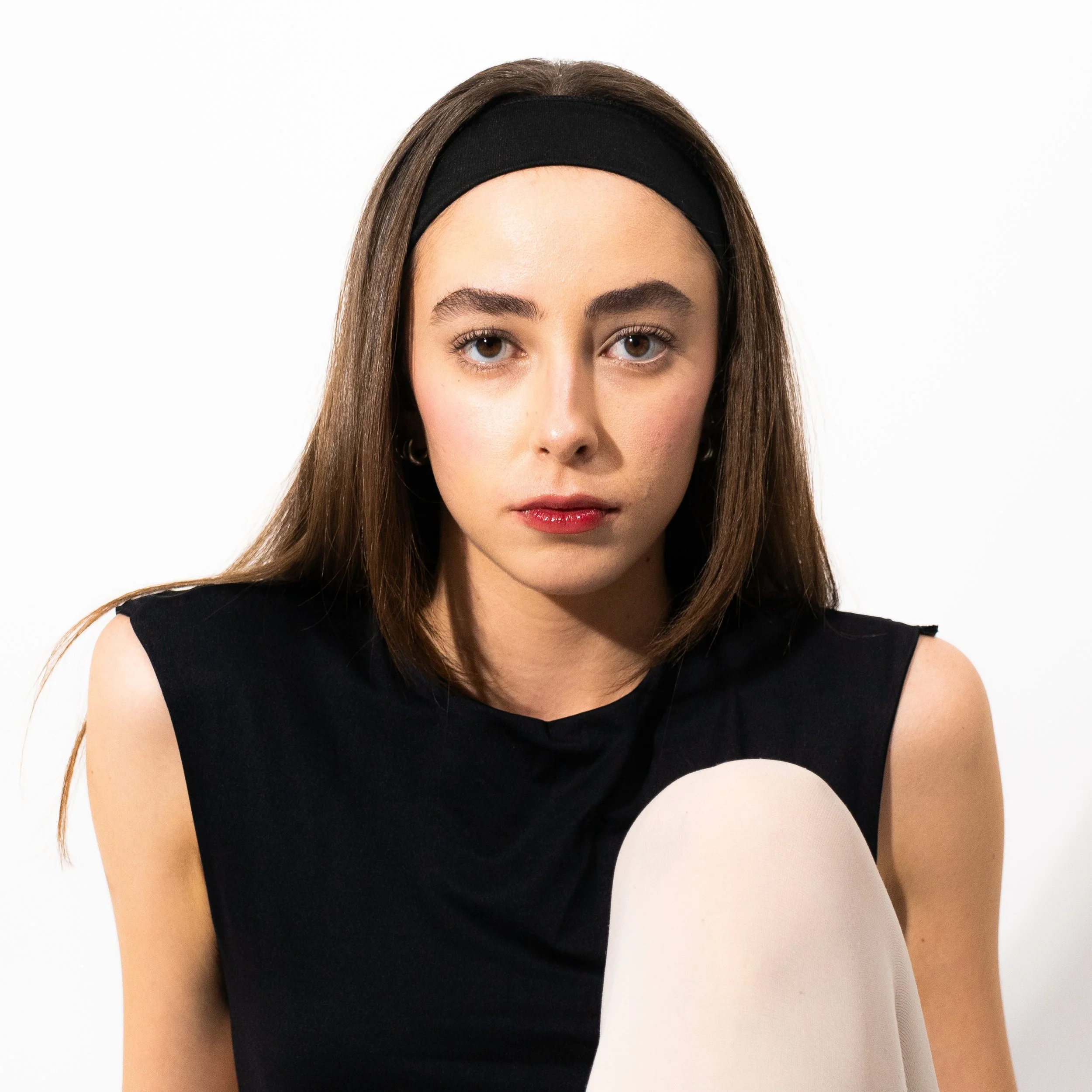 Young woman with long brown hair, wearing a black sleeveless top and headband, sitting against a white background.