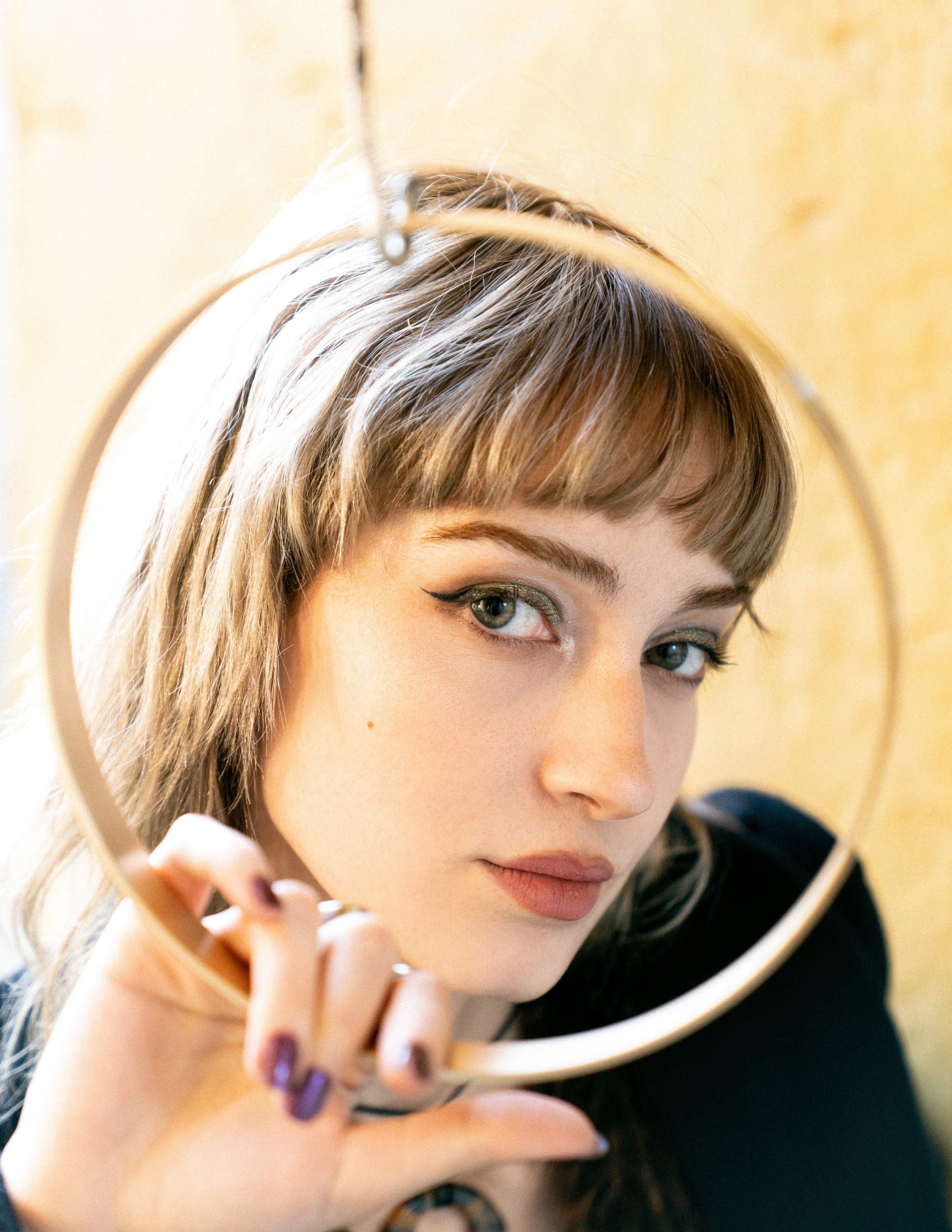 A young woman with blonde hair and makeup, holding a round mirror up to her face and looking into it.