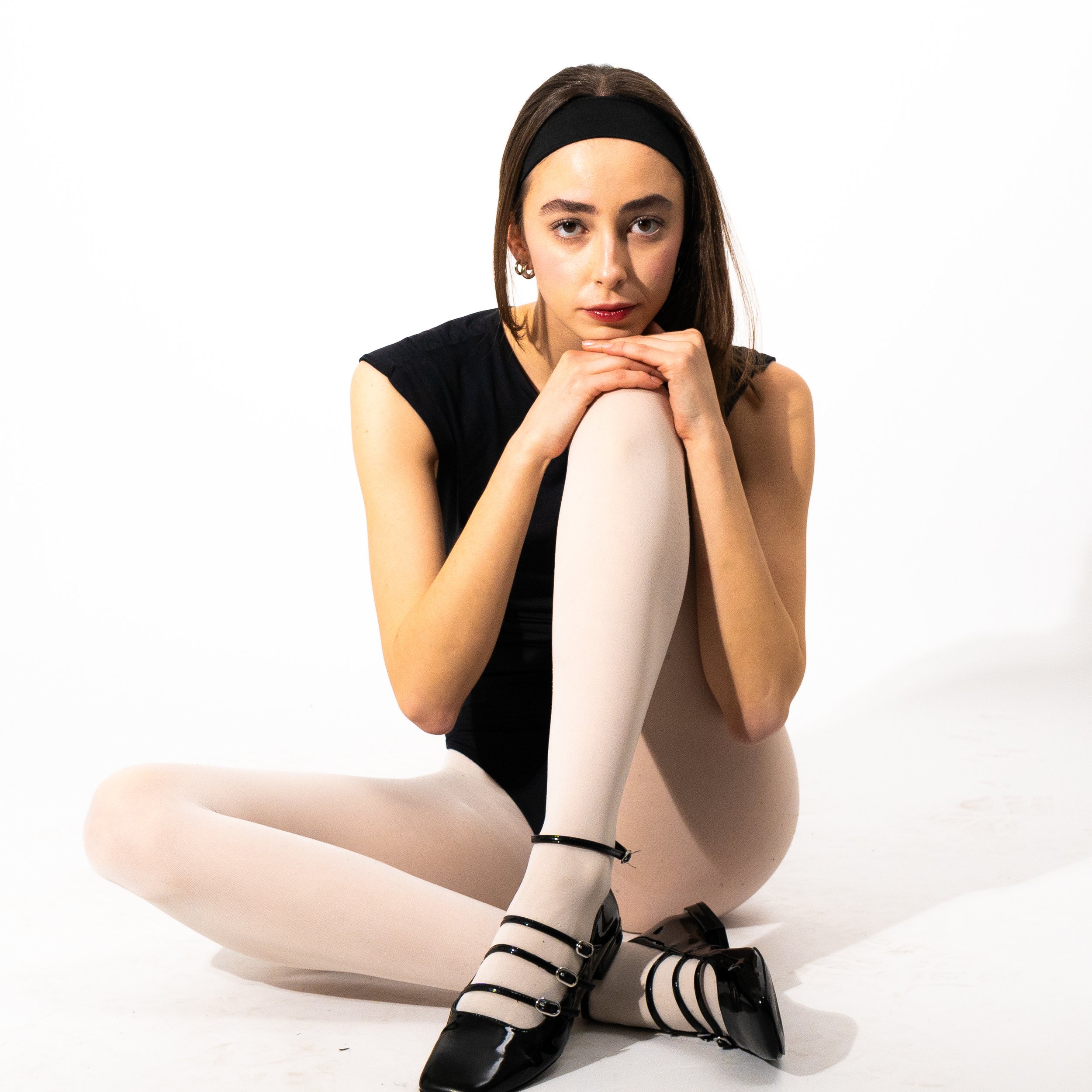Young woman sitting on the floor with one leg over the other, resting her chin on her knee, wearing black sleeveless top, white tights, black shoes with straps, and a black headband, against a white background.