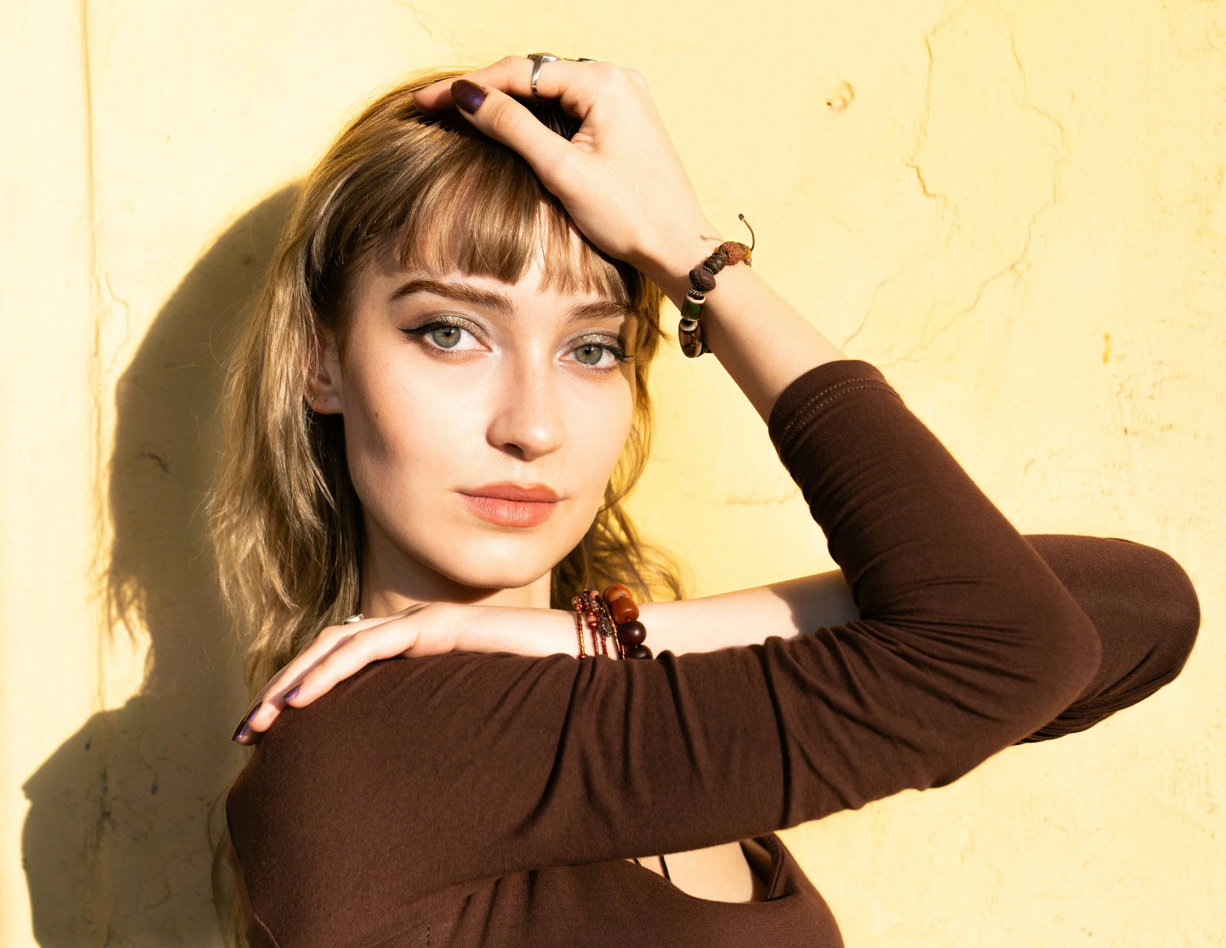 Young woman with light brown hair and green eyes posing against a yellow wall, wearing a brown top and colorful bracelets, with her hand on her forehead and the other resting on her shoulder.