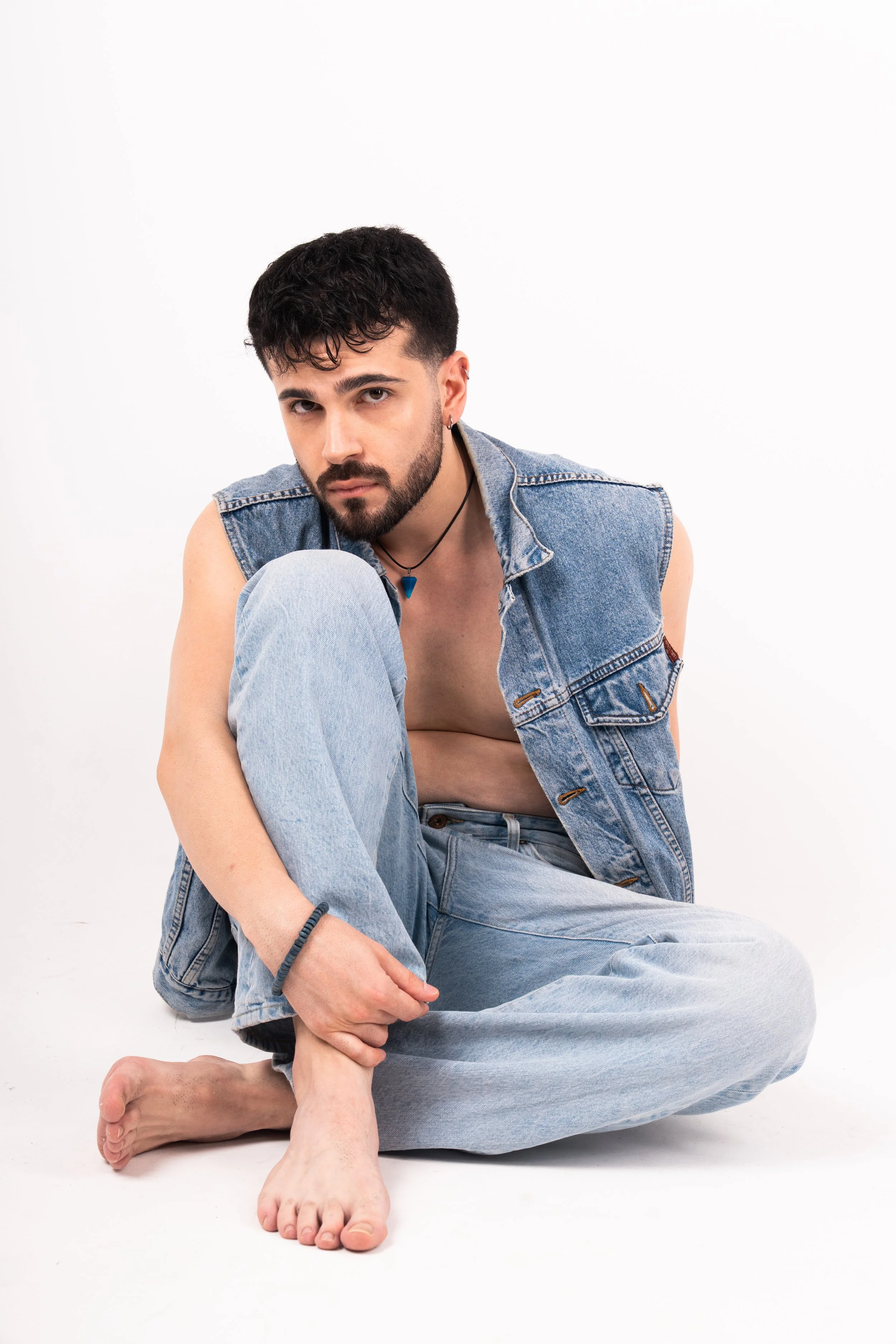 A young man with dark hair, beard, and earrings sitting on the floor against a white background. He is wearing a sleeveless denim vest and light-wash jeans, with one leg crossed and his arm resting on his knee. He looks directly at the camera.
