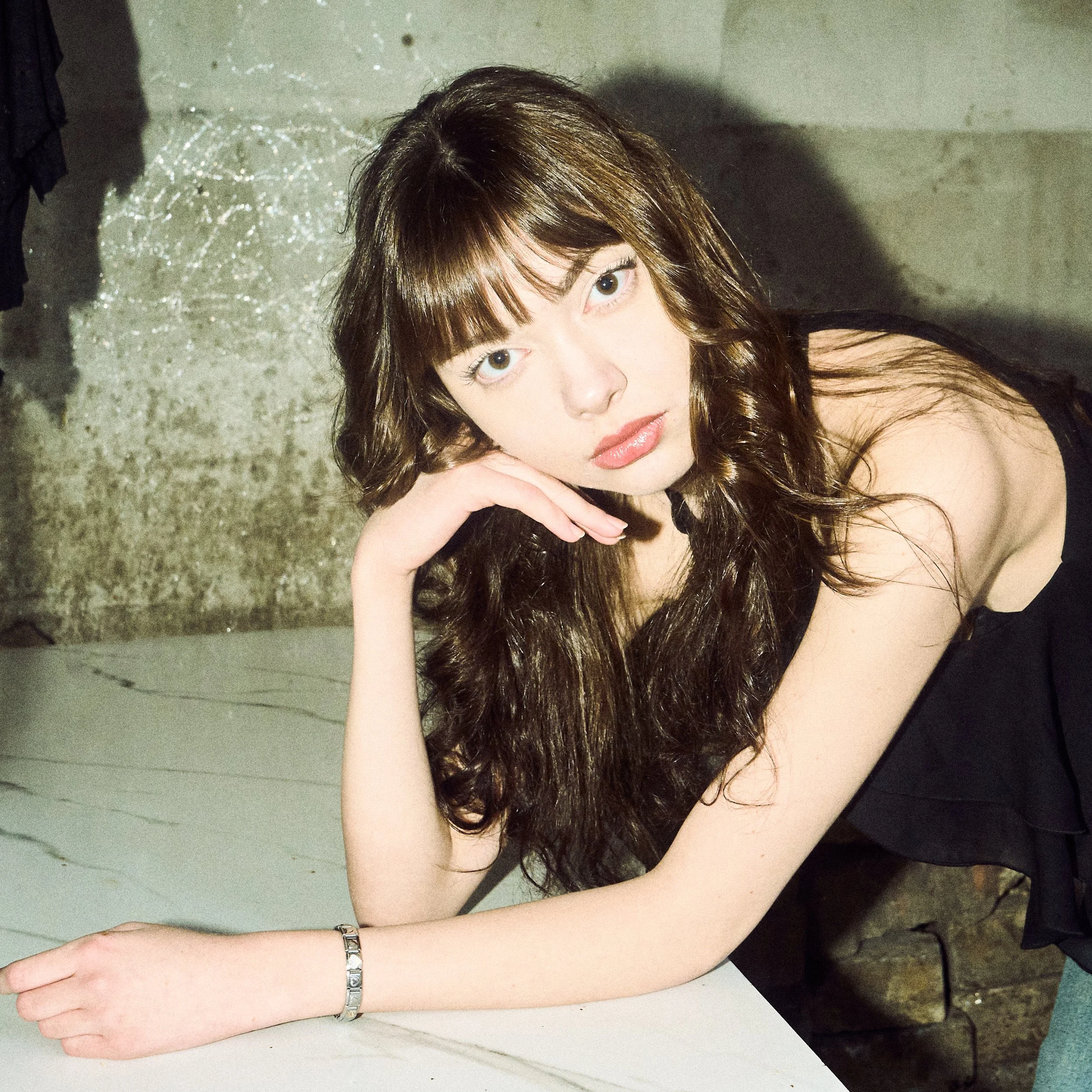 A young woman with curly brown hair and light skin, resting her head on her hand, leaning on a white marble table in an industrial setting with a concrete wall in the background, wearing a black off-shoulder top and a silver bracelet.