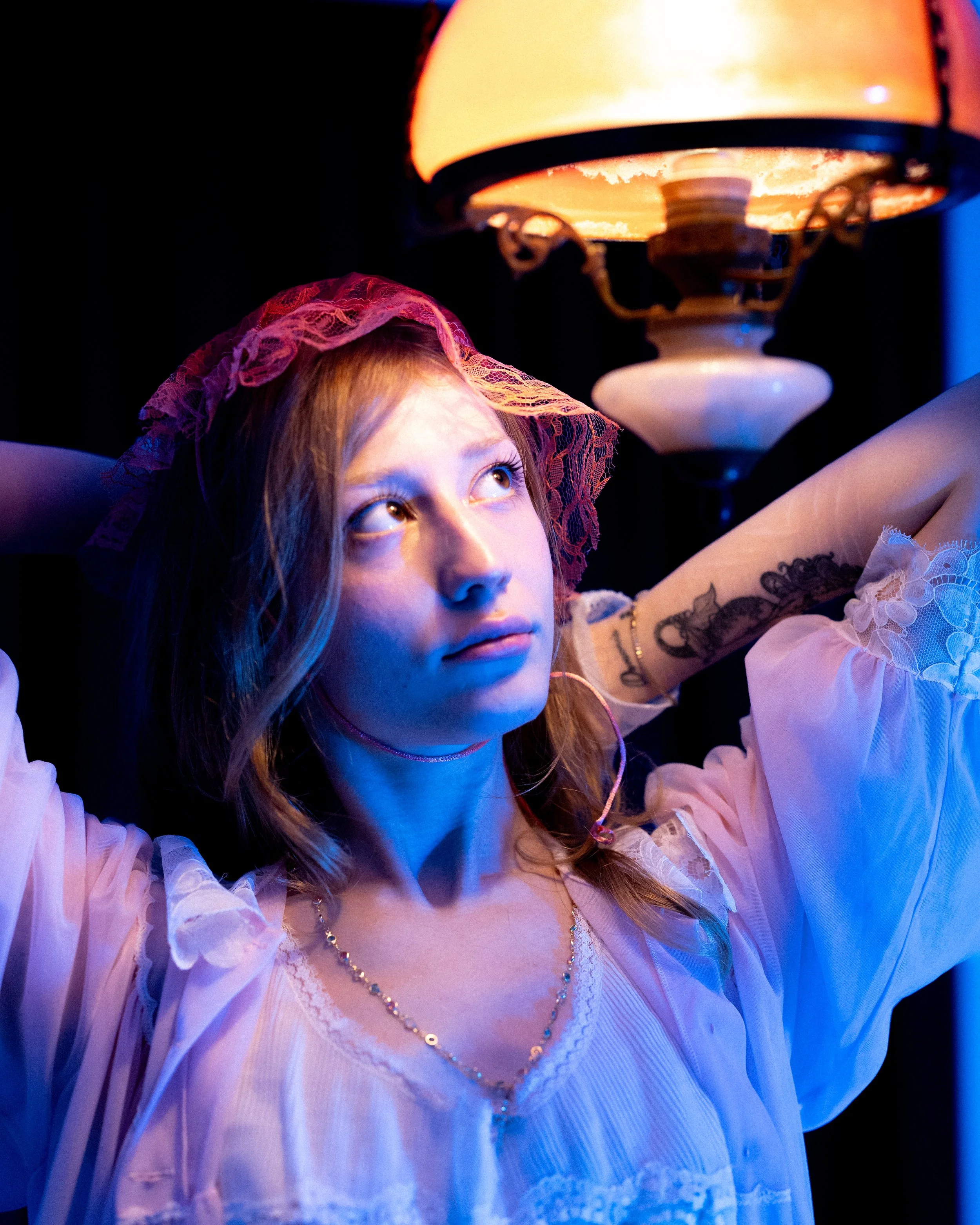 A woman with wavy hair wearing a pink lace headscarf and a white blouse, illuminated by a vintage orange lampshade, looks upward with a thoughtful expression.