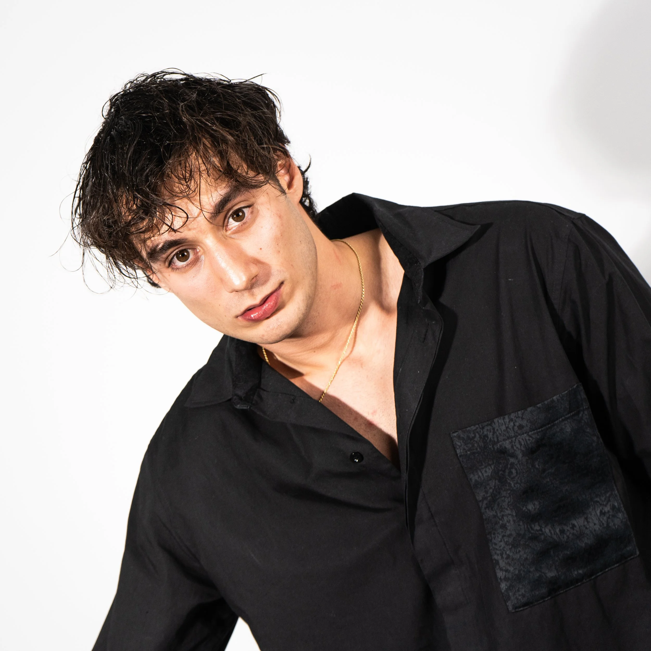 A young man with brown, curly hair and light skin wearing a black shirt with a shiny black pocket and a gold chain necklace, posing against a plain white background.