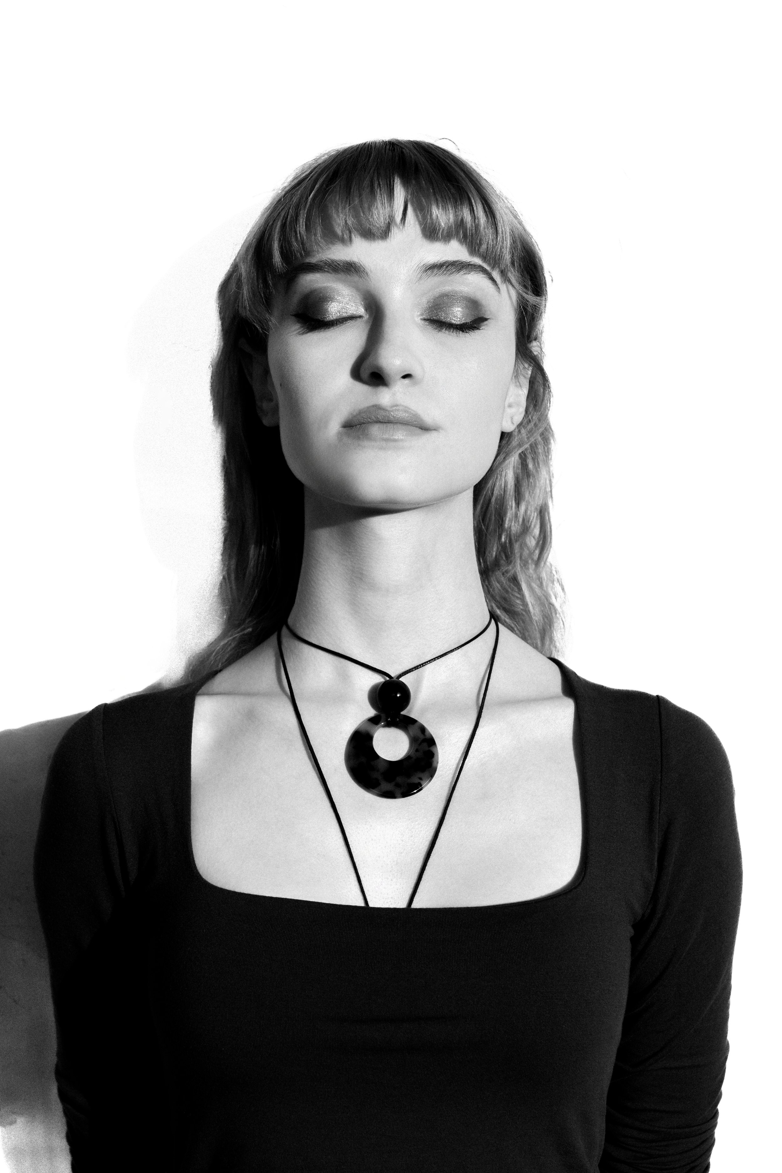 Black and white portrait of a woman with eyes closed, wearing layered necklaces with geometric pendants, and a black top with a square neckline.