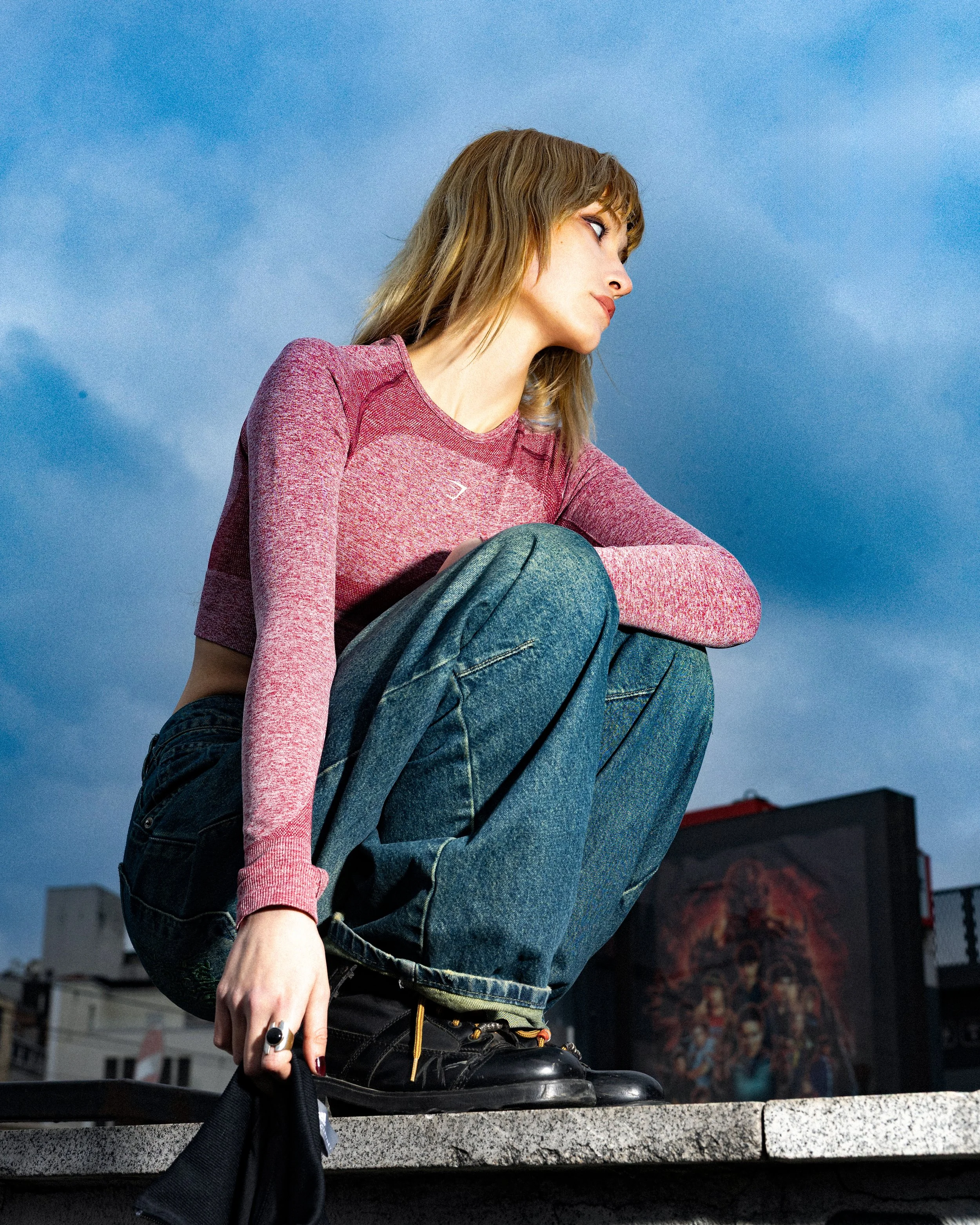 A woman with shoulder-length hair wearing a pink long-sleeve shirt, jeans, and boots, squatting on a ledge with a cityscape and a cloudy sky in the background.