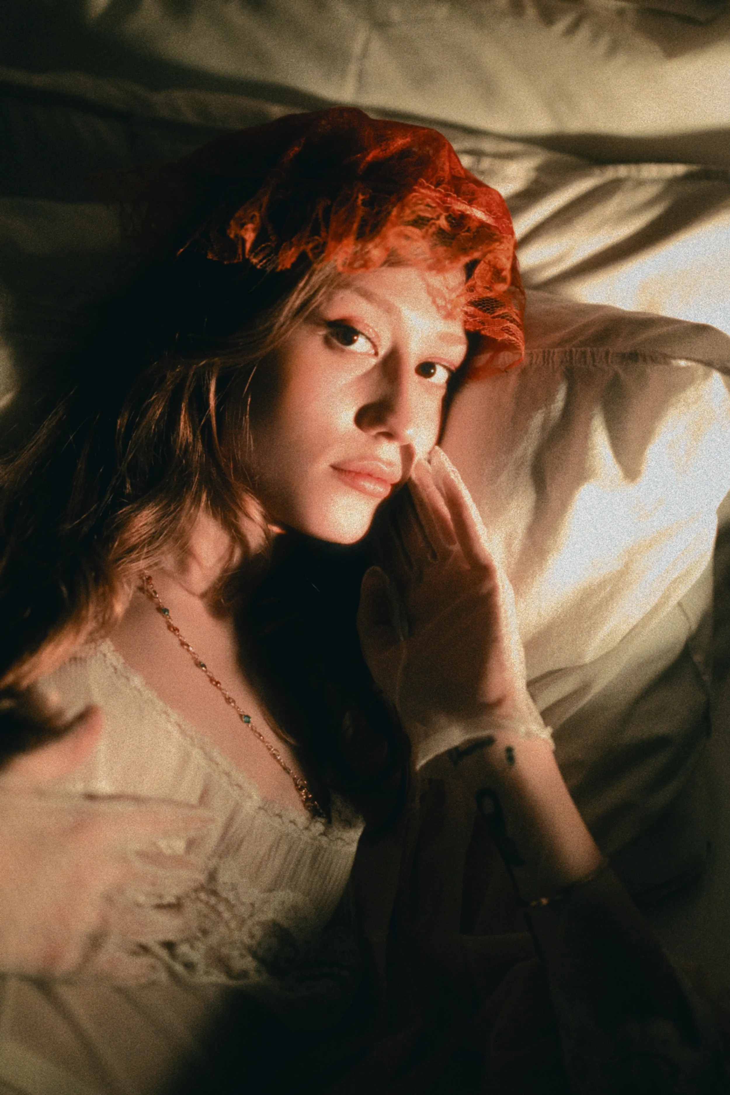 A young woman with long wavy hair lying on a bed, wearing a red lace headscarf and a light-colored top, looking directly at the camera with a gentle expression, illuminated by warm, soft lighting.