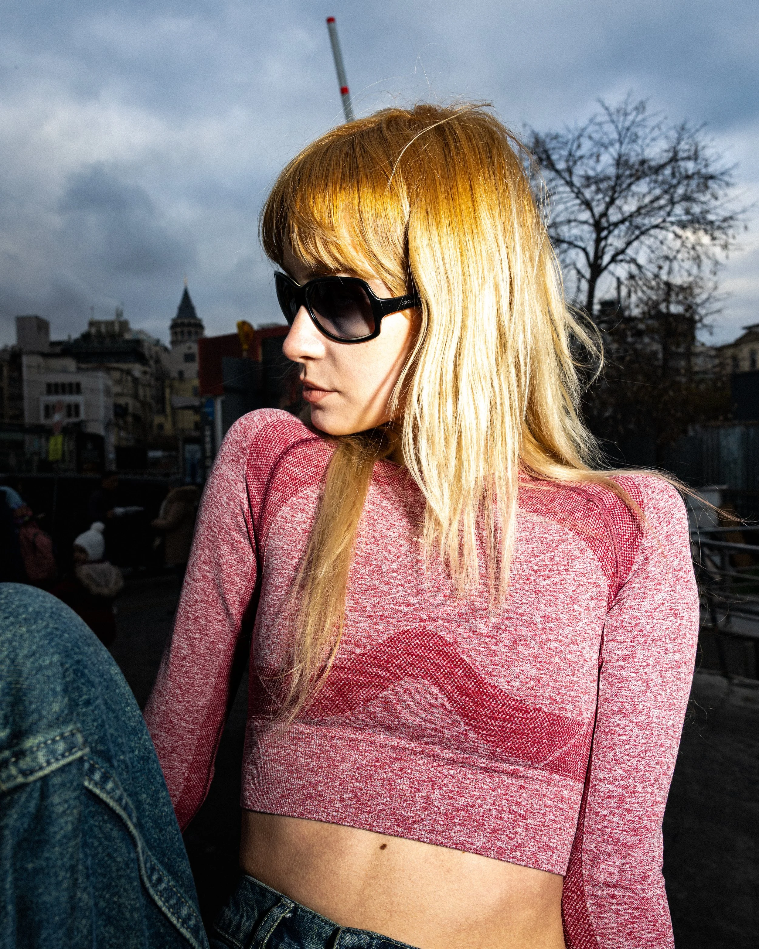 A woman with blonde hair wearing black sunglasses and a pink long-sleeve crop top, sitting outdoors on a cloudy day with city buildings in the background.