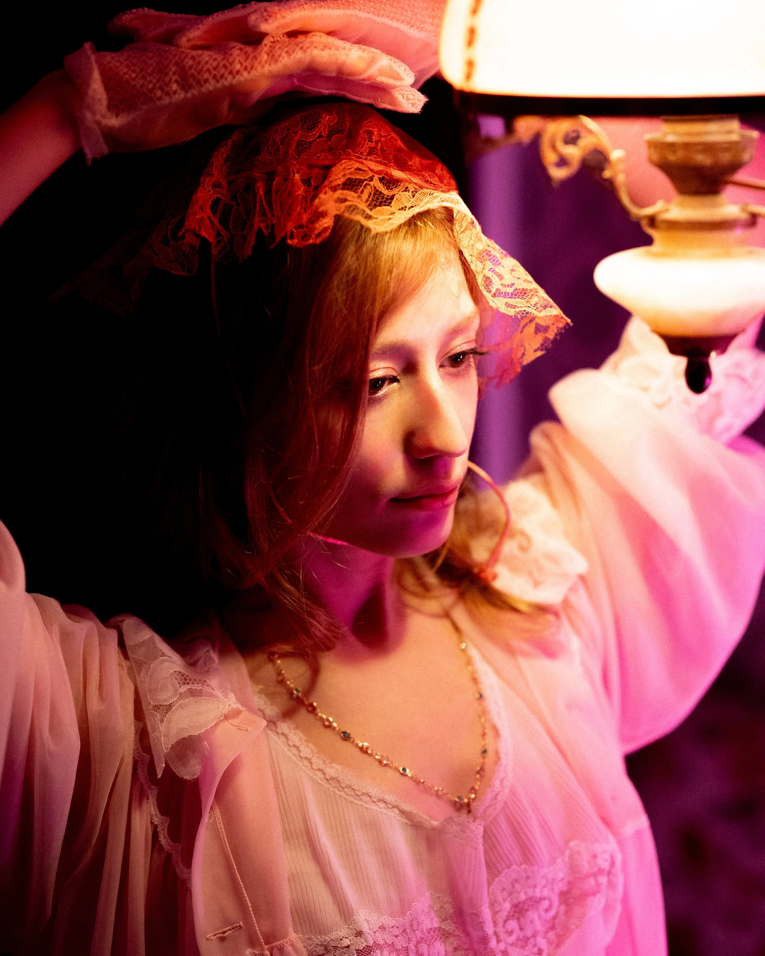 A young woman with red hair in vintage pink nightgown and lace bonnet, standing next to a lit vintage lamp, with her hand resting on her head, looking down thoughtfully.
