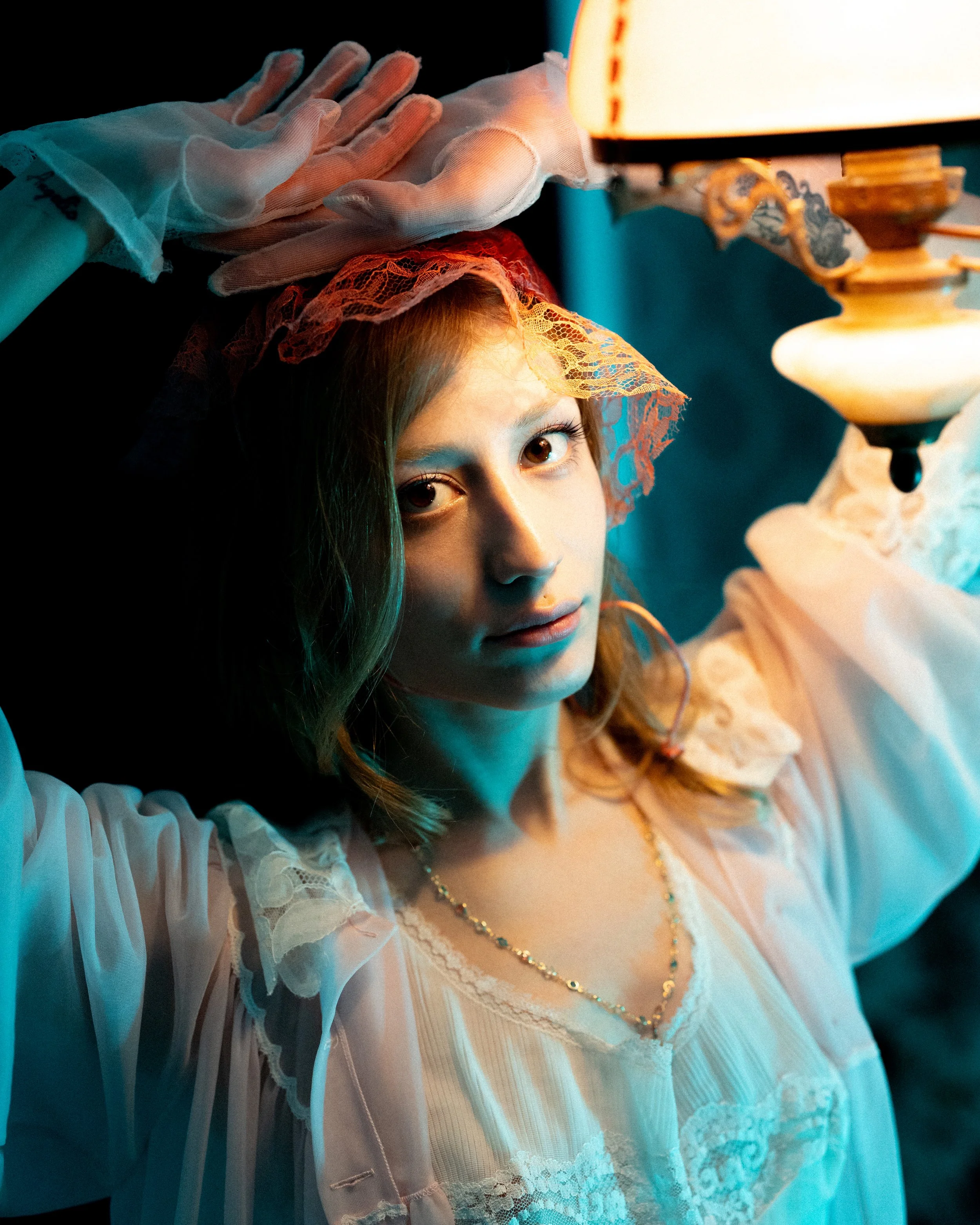 A woman with red hair in vintage clothing, wearing lace gloves and a lace headpiece, sitting under a warm light with a serious expression.
