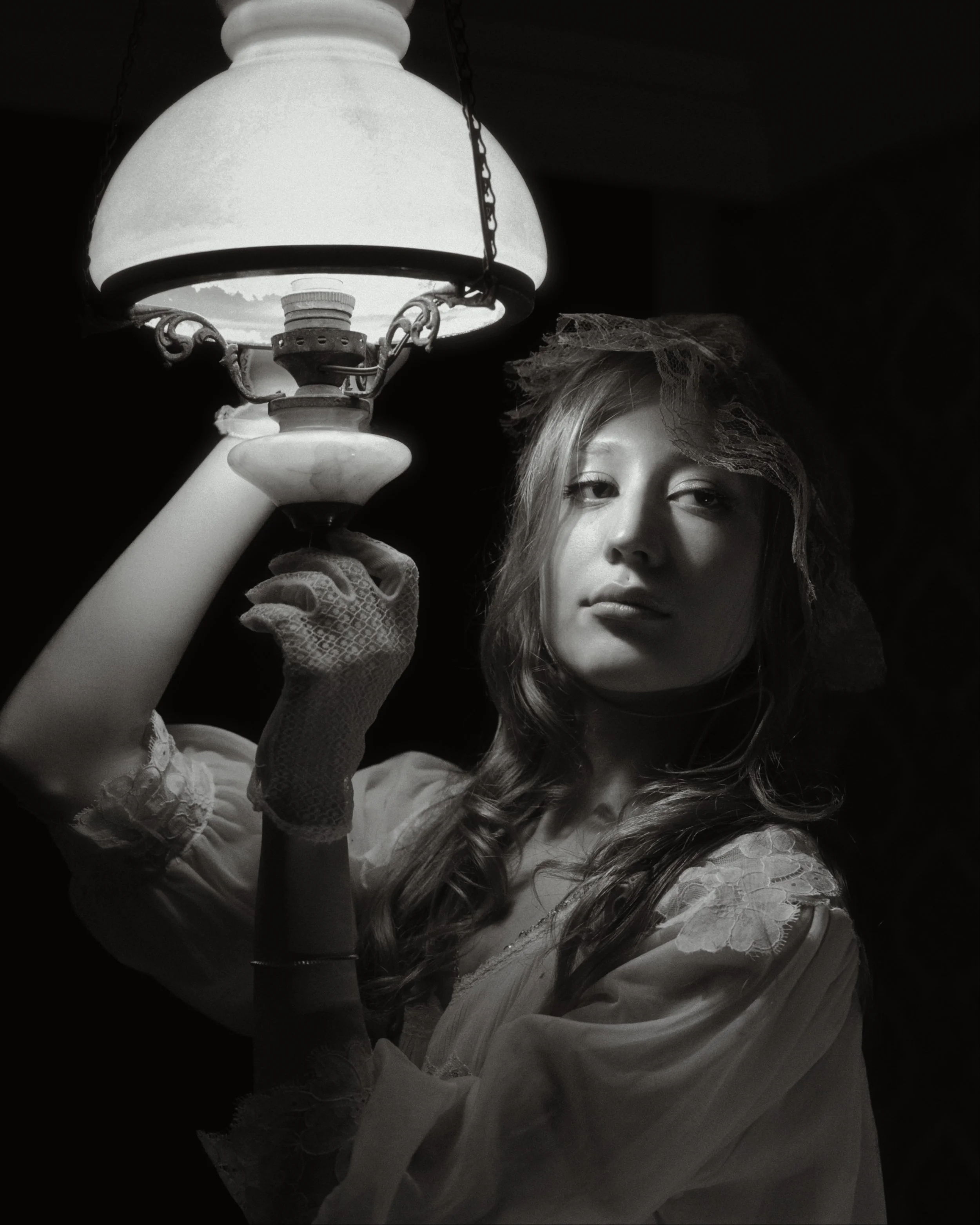 A young woman with wavy hair wearing lace gloves is holding an antique-style lamp and looking into the camera in a black and white portrait.