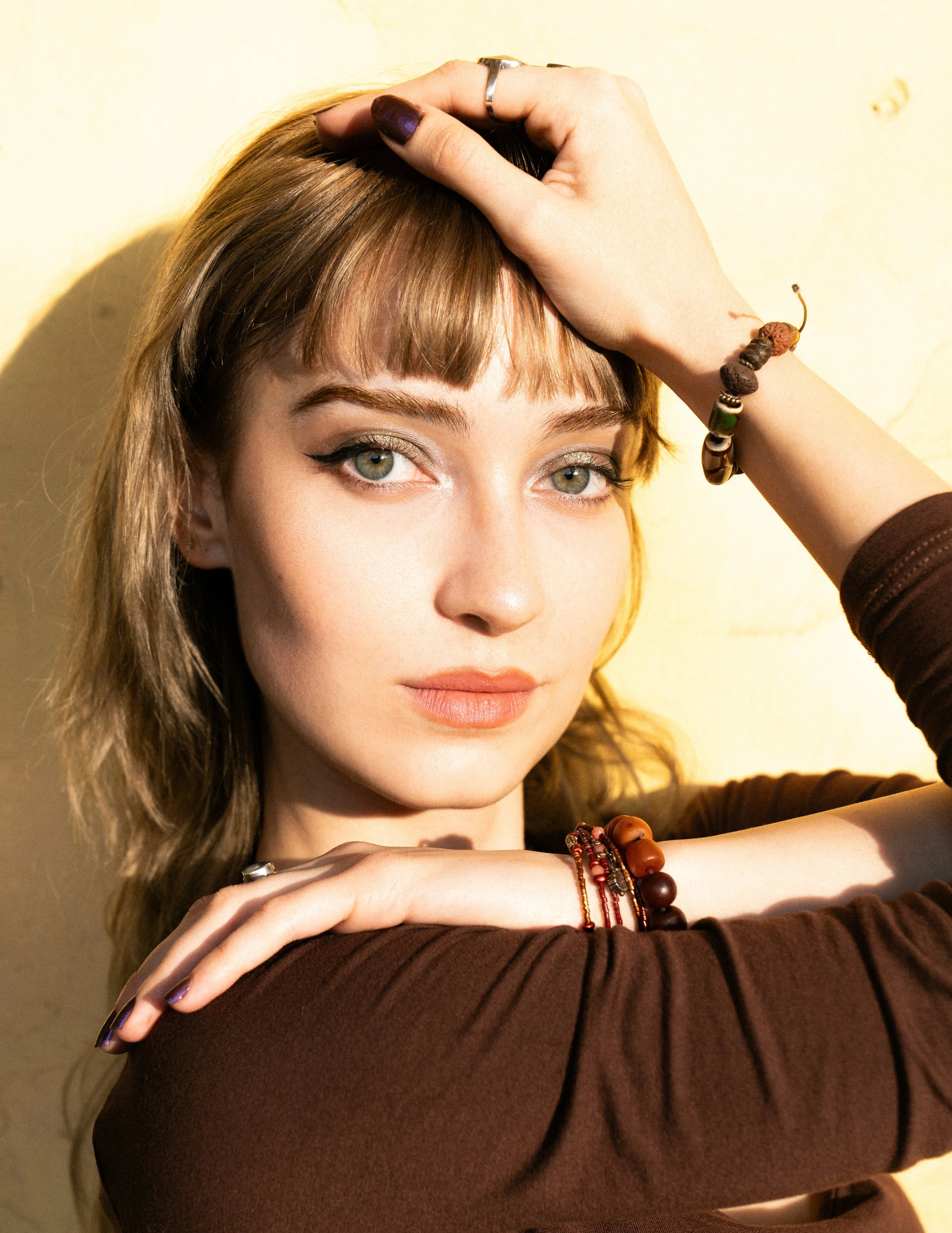A young woman with light brown hair, green eyes, and natural makeup, resting her head on her arm while touching her forehead with her hand, wearing multiple bracelets and a brown long-sleeve shirt, standing against a beige wall.