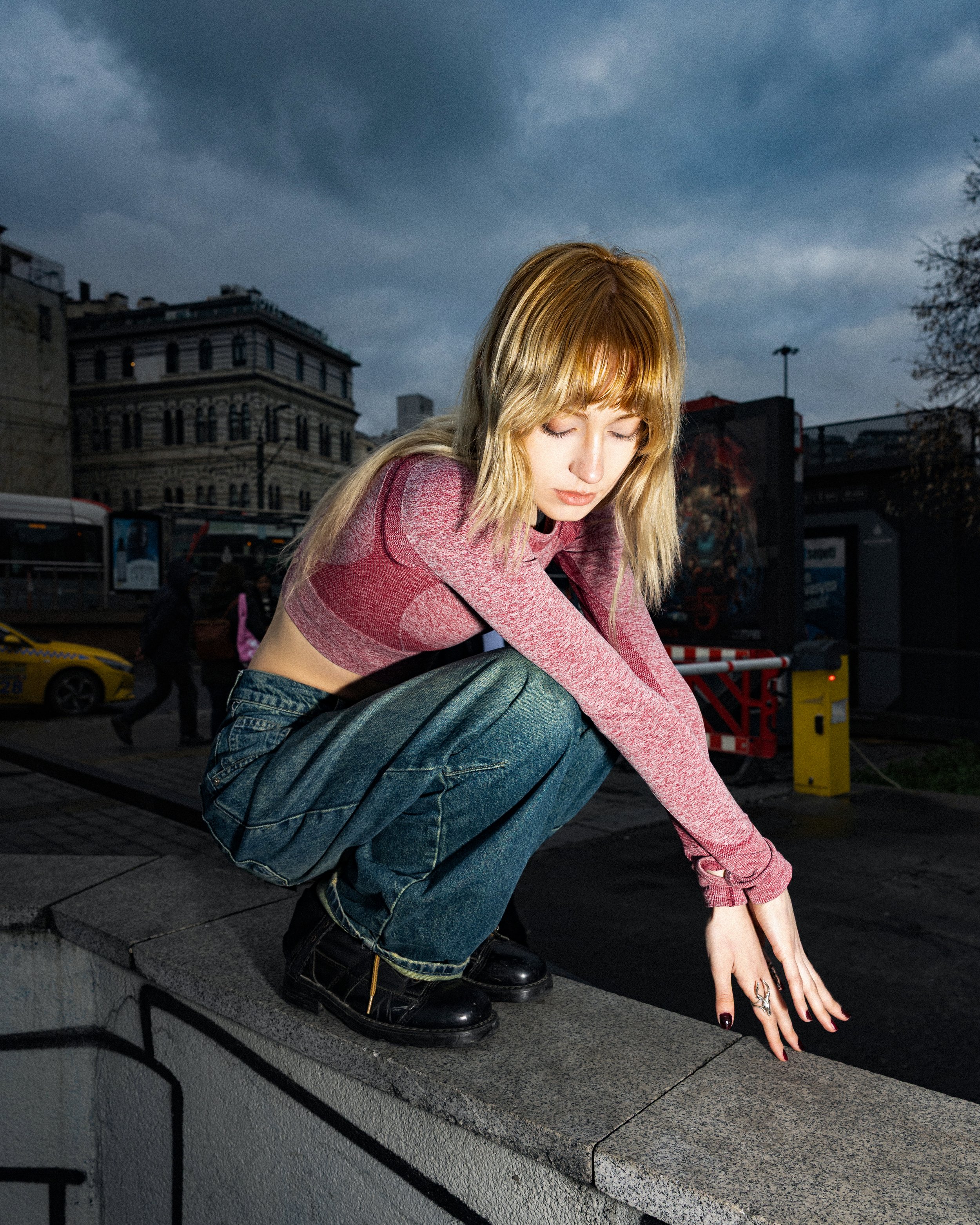 A young woman with blonde hair squatting on a city ledge during dusk, wearing a pink long-sleeve crop top, loose jeans, and black boots, with her hands touching the ledge and eyes closed against a cloudy sky.